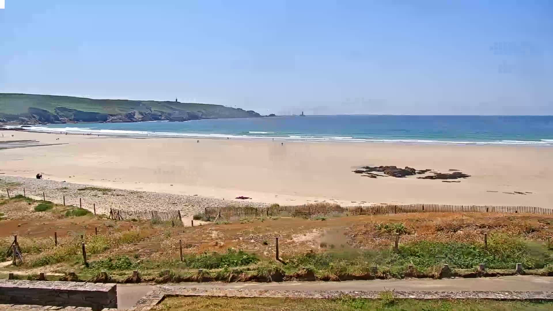 A sandy beach with small waves and a few people stretches out to a calm ocean under a clear blue sky, backed by green hills.