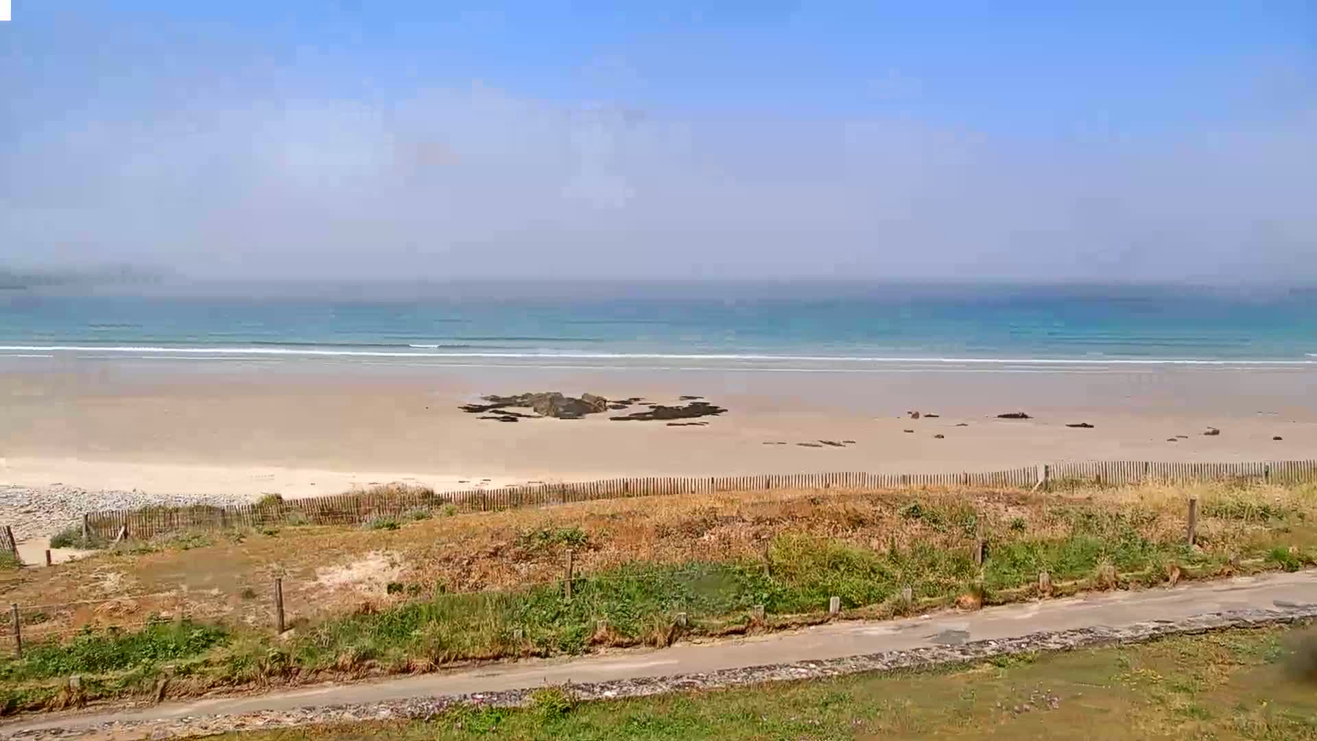 A sandy beach with small waves on a hazy, sunny day is seen from an elevated vantage point beyond a wooden fence and a road.