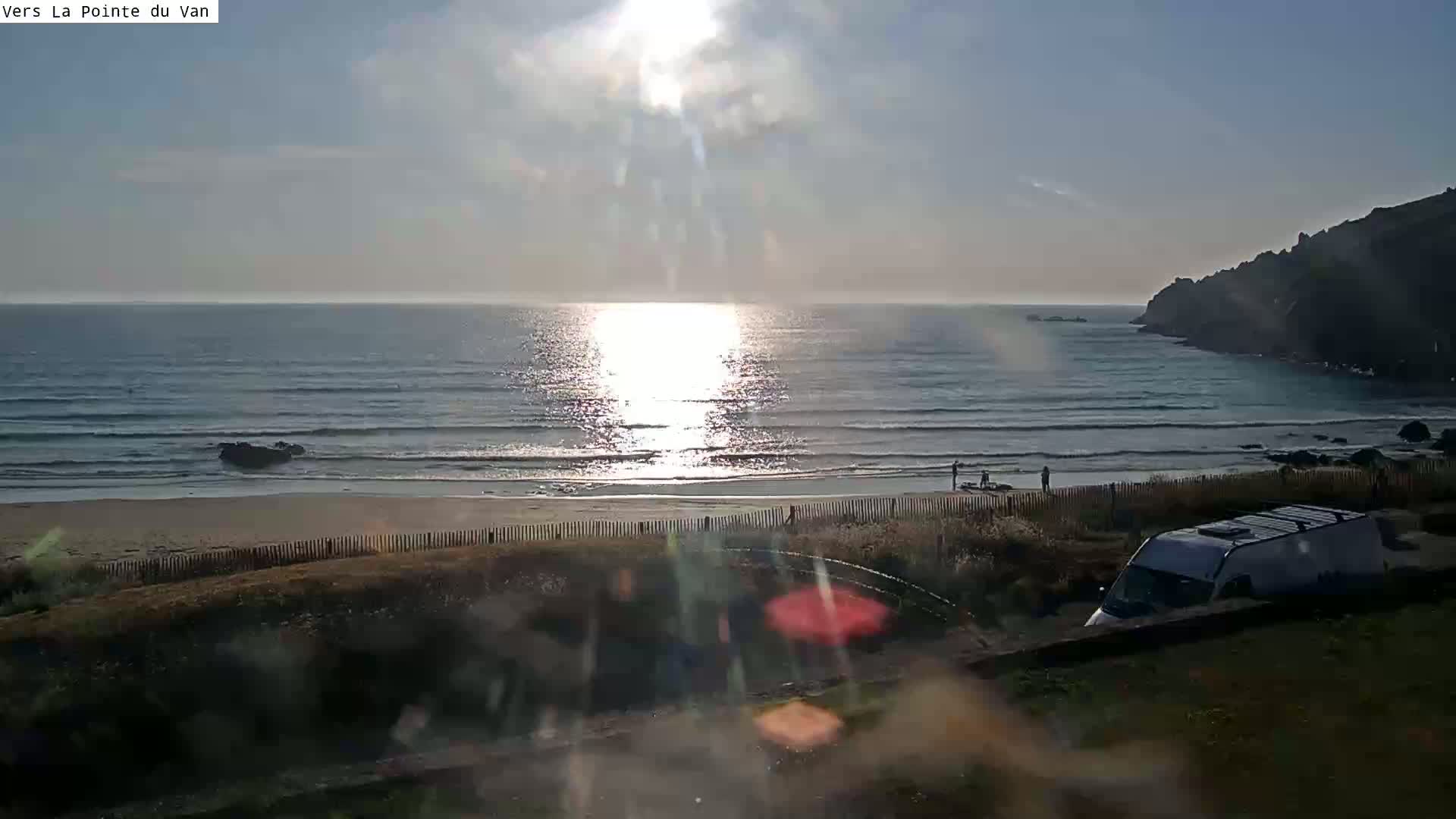 A sandy beach with gentle waves and a bright sun reflecting on the ocean is seen from a distance, with a van parked nearby on a grassy area.