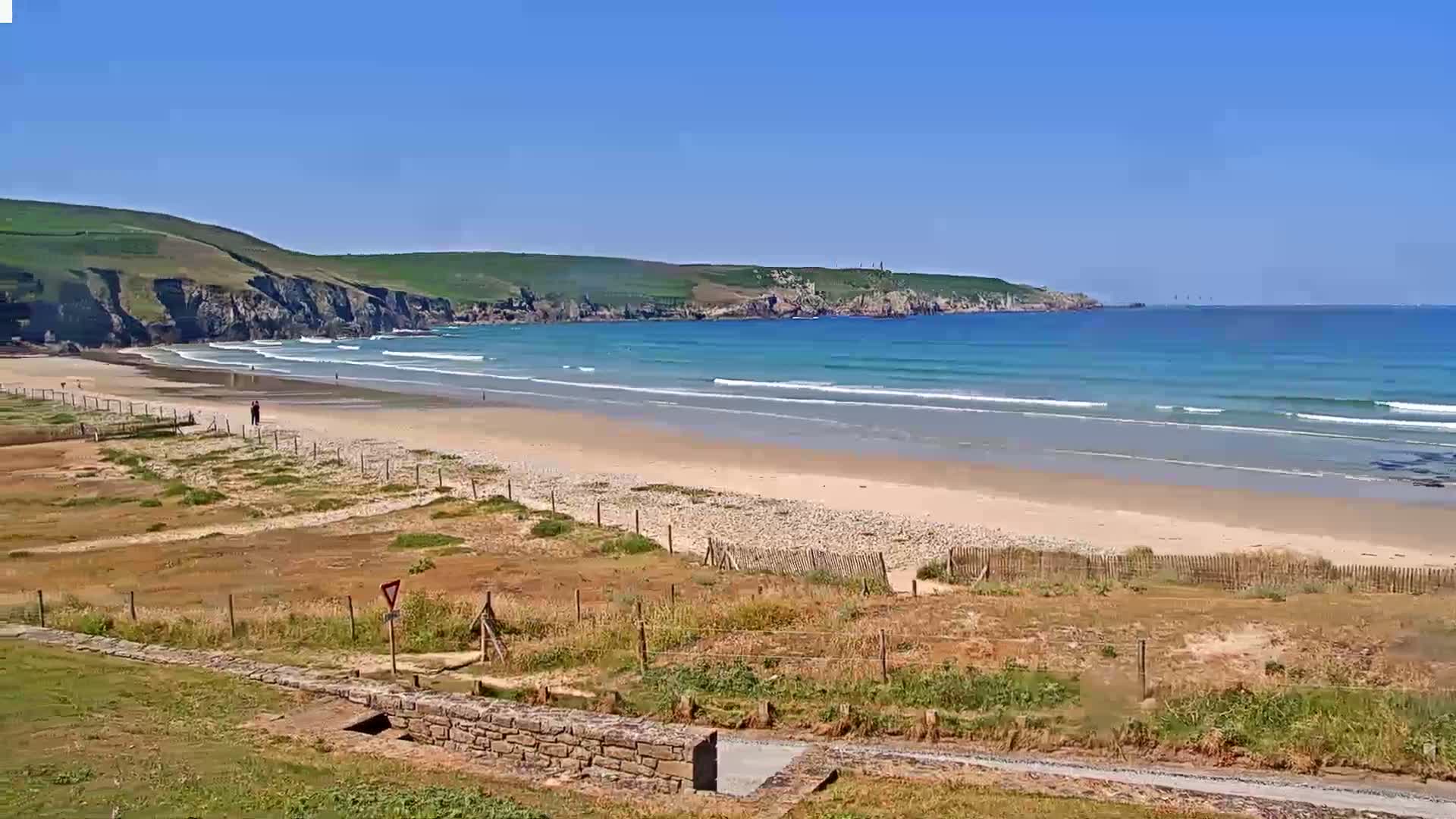 A sandy beach with gentle waves under a clear blue sky stretches along a coastline backed by green hills.