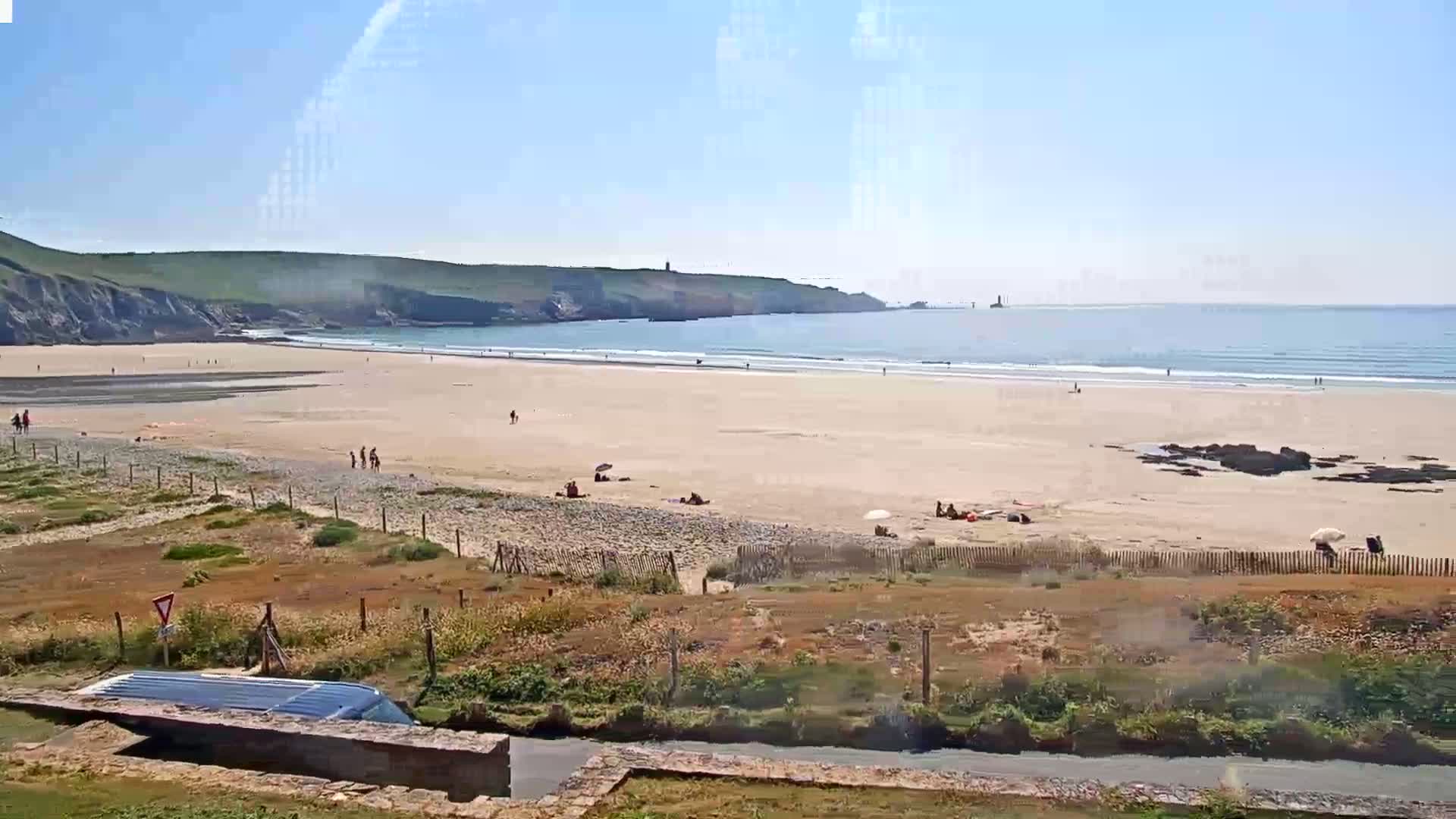 A sandy beach stretches along a calm ocean under a clear blue sky, with a few people scattered across the sand and a vehicle partially obscured by a low wall in the foreground.