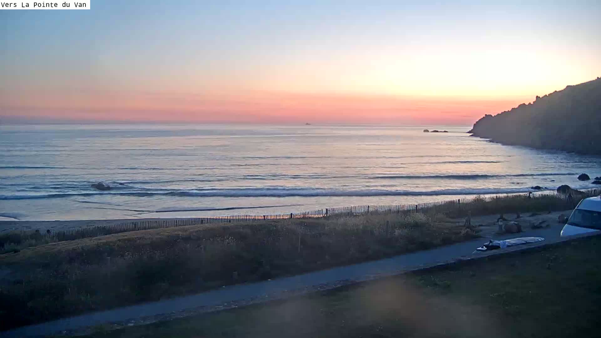 A pink and orange sunset over a calm ocean with small waves lapping a sandy beach behind a fence, observed from a vantage point above.