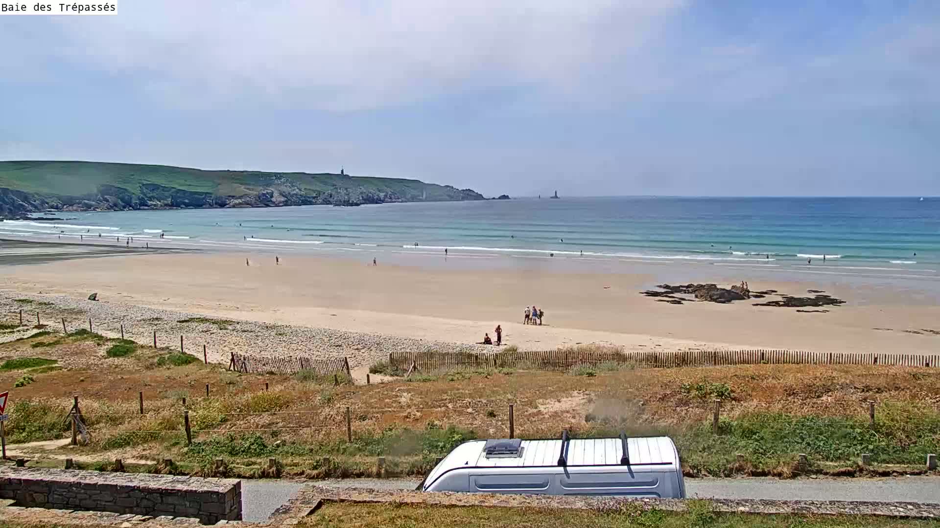 A sandy beach with a few people on it stretches out to a calm ocean under a mostly sunny sky, with a camper van partially visible in the foreground.