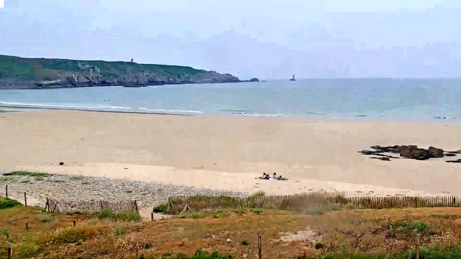 A sandy beach stretches out to a calm, light blue ocean under a mostly cloudy sky, with a few people relaxing on the sand and a distant rocky headland.