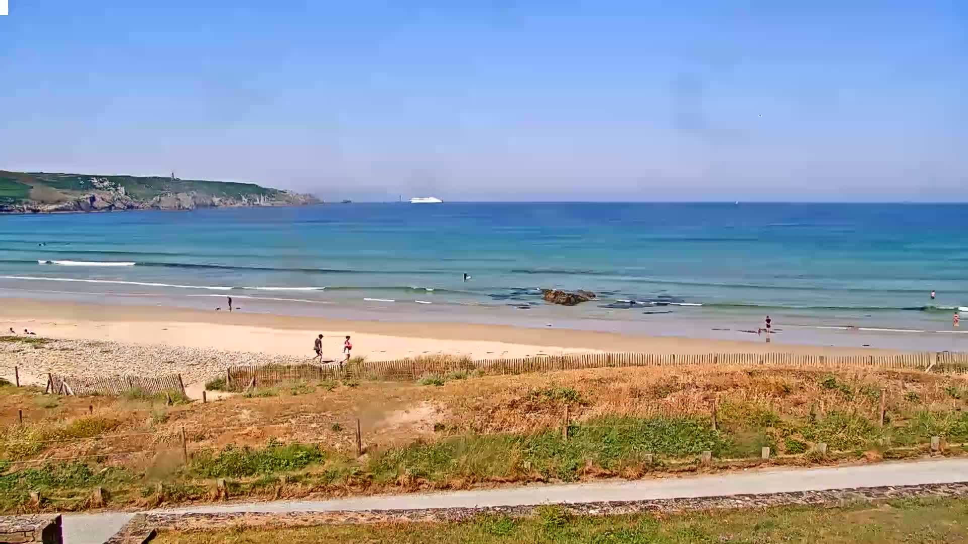 A sandy beach with gentle waves under a clear blue sky, a few people are seen on the beach and a large ship is visible on the horizon.