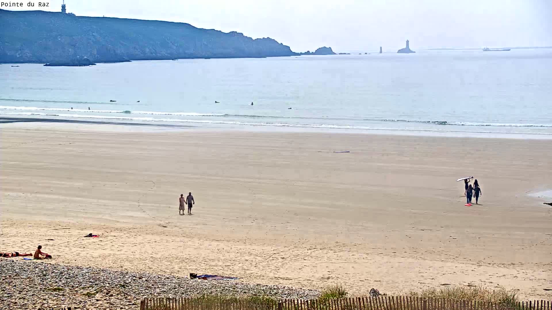 A sandy beach with a few people sunbathing and walking, some surfers in the ocean, and a distant coastline with a lighthouse under a hazy sky.