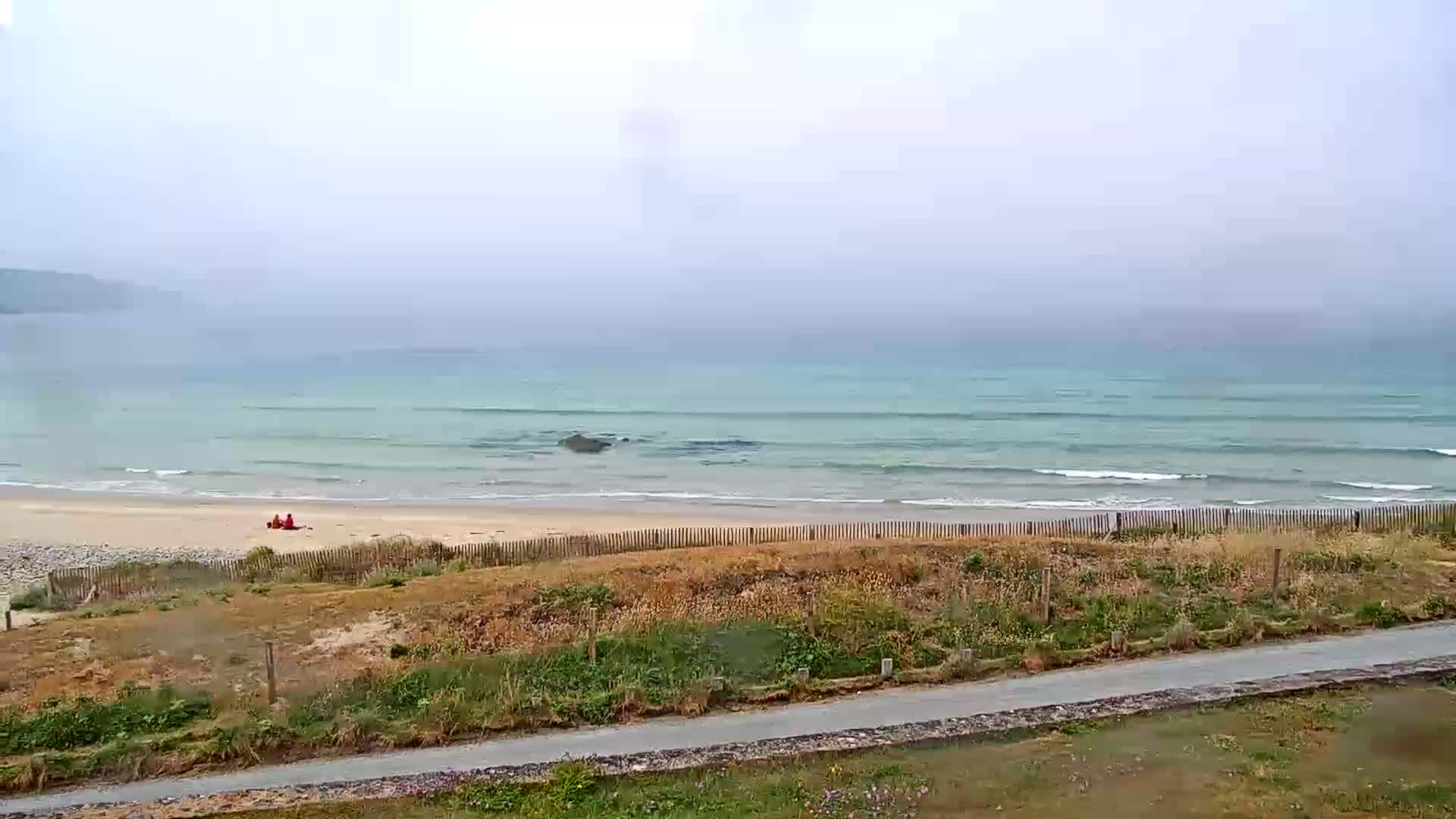 A hazy view shows a sandy beach with small waves lapping the shore, behind a dune and wooden fence, with two figures sitting on the sand.