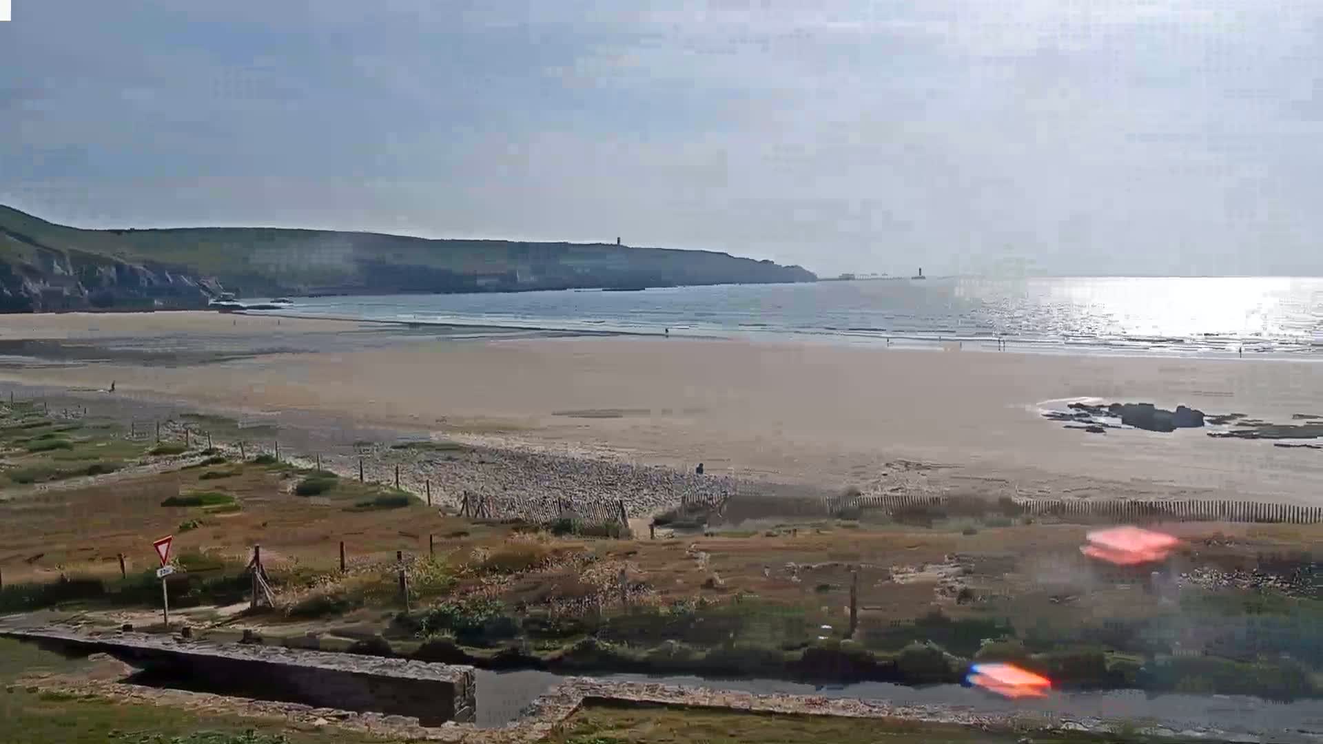 A sandy beach stretches to a calm ocean under a bright, sunny sky, with a small stream and grassy dunes in the foreground.