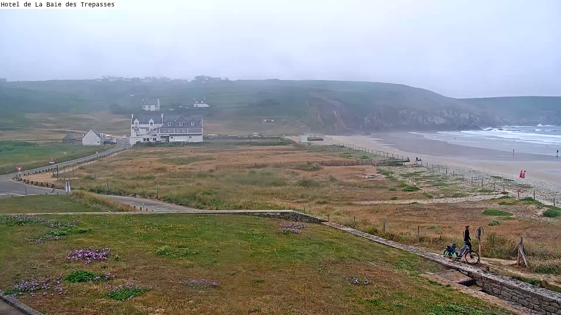 A person on a bicycle is seen near a sandy beach, with a hotel and other buildings visible in the distance under a foggy sky.