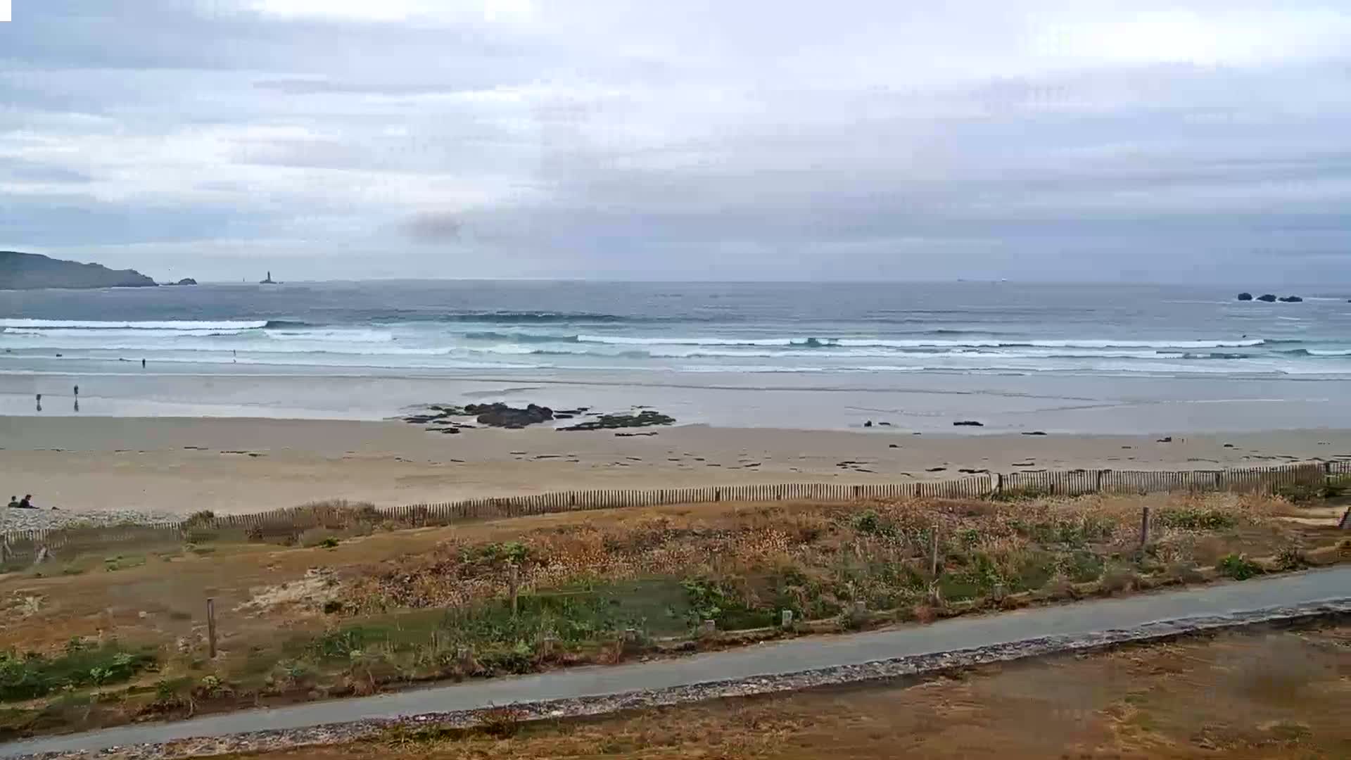A sandy beach with gentle waves under a cloudy sky is viewed from above a low-lying area with sparse vegetation.