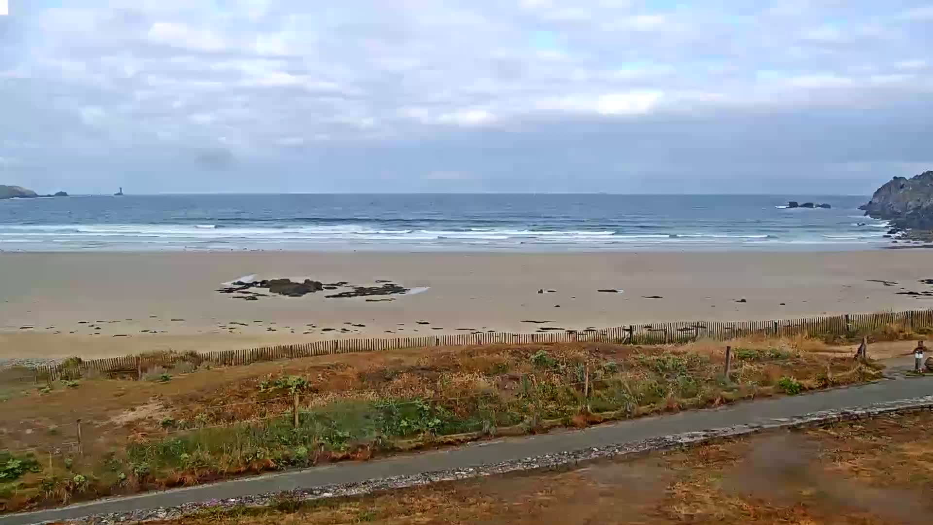 A sandy beach with small waves under a mostly cloudy sky is viewed from a grassy area above a paved path.