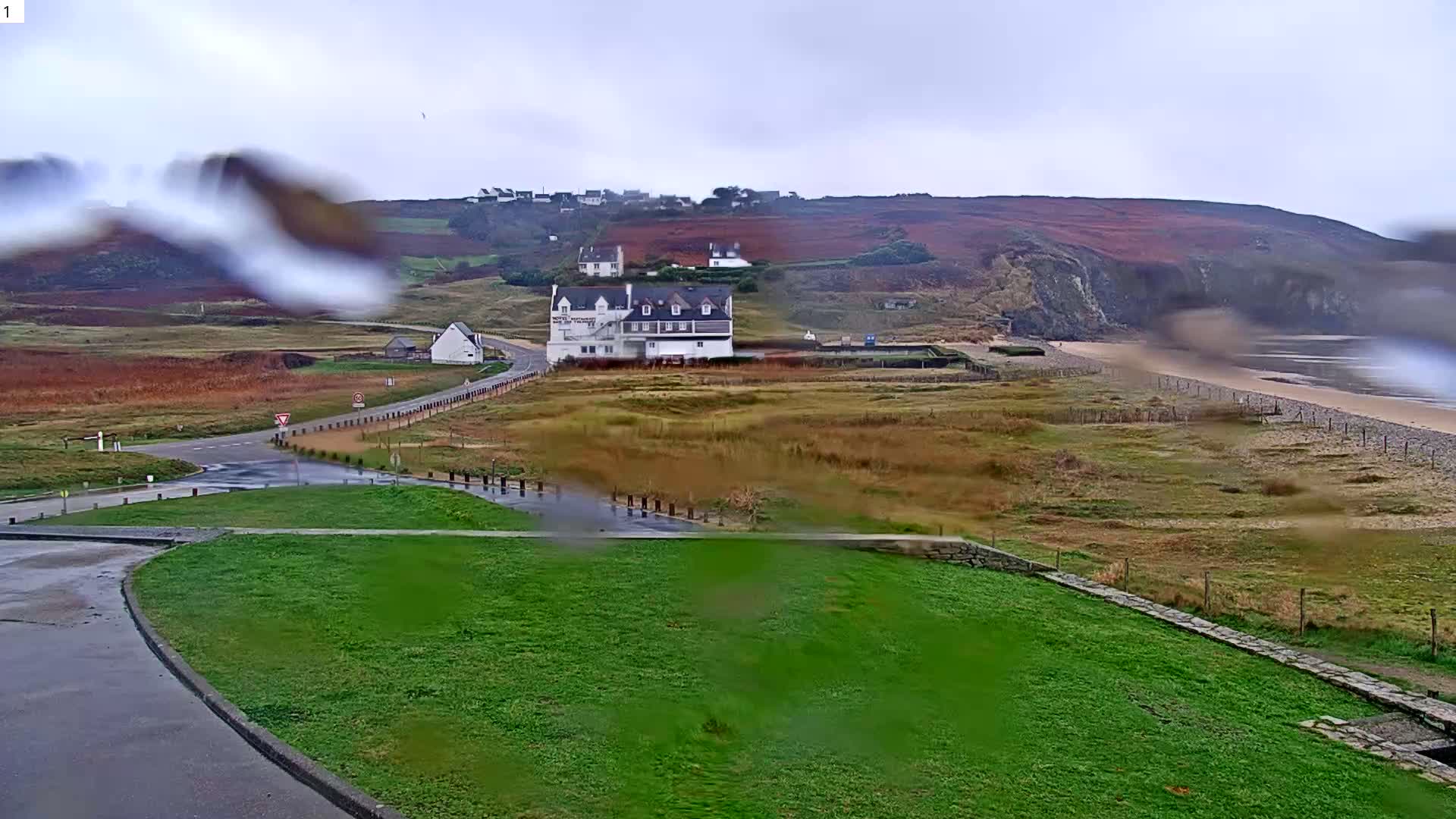 On an overcast and rainy day, a coastal landscape unfolds with a scattering of white buildings amidst reddish-brown hills, winding wet roads, green fields in the foreground, and a sandy beach stretching towards the sea under a grey sky.
