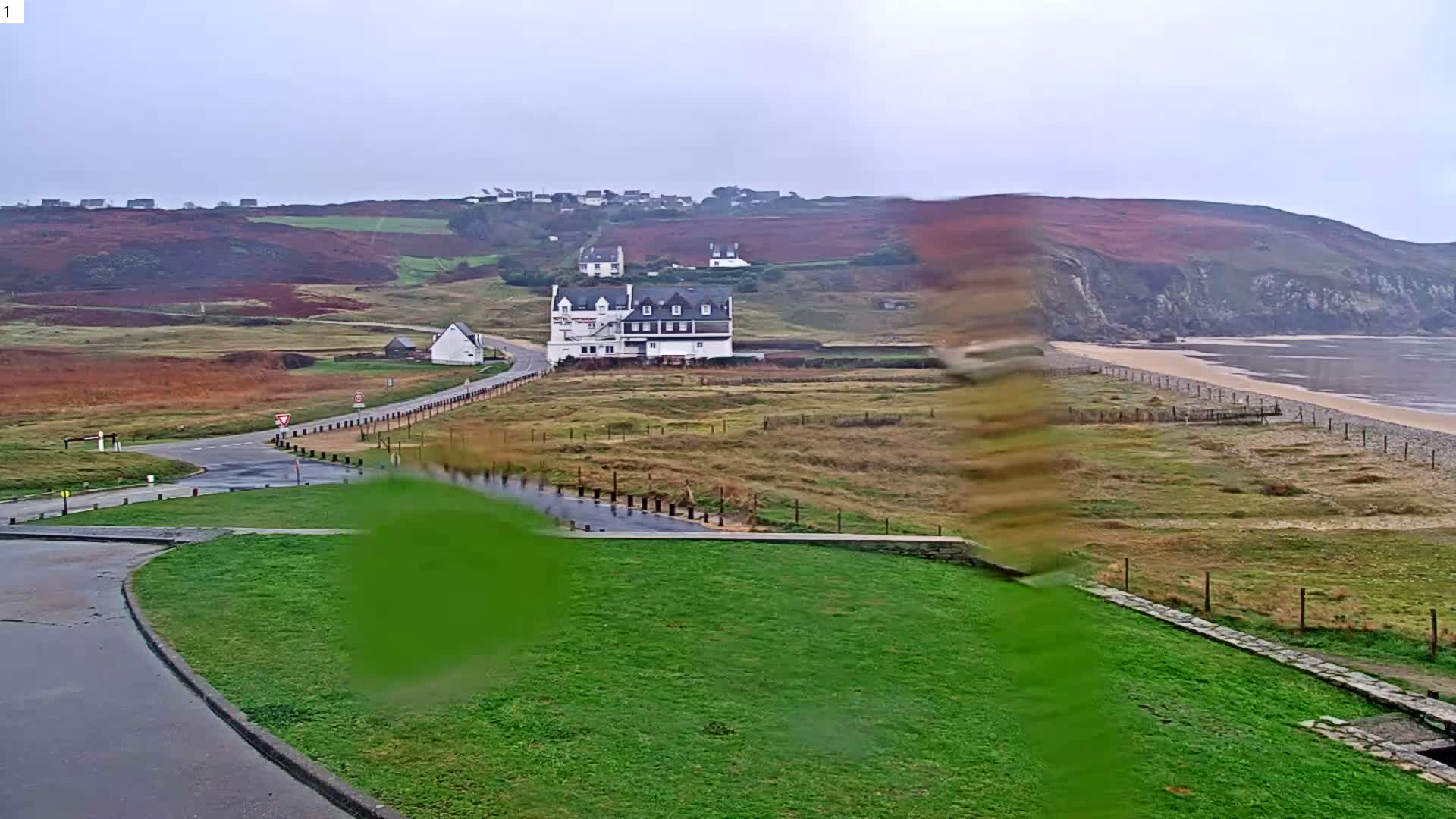 A wide coastal landscape on an overcast and damp day showcases a winding road, green and reddish-brown fields, a large white building, and distant houses on rolling hills, all leading towards a pebbled beach and the calm sea beneath grey skies.