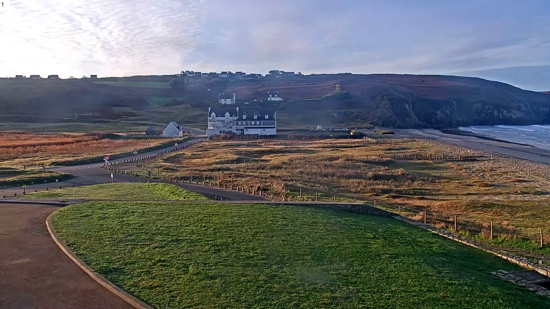 The image shows a bright, clear coastal landscape on a sunny day, featuring a large white building near a road intersection surrounded by fields of dry grass and fences, with a village on distant hills, and a sandy beach with ocean waves at the foot of rugged cliffs.