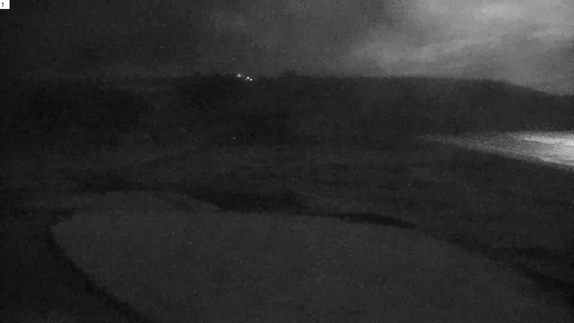 A dark, cloudy night scene reveals a wide expanse of land or beach in the foreground, leading to a body of water reflecting light on the right, with faint lights visible on a distant, hilly horizon.