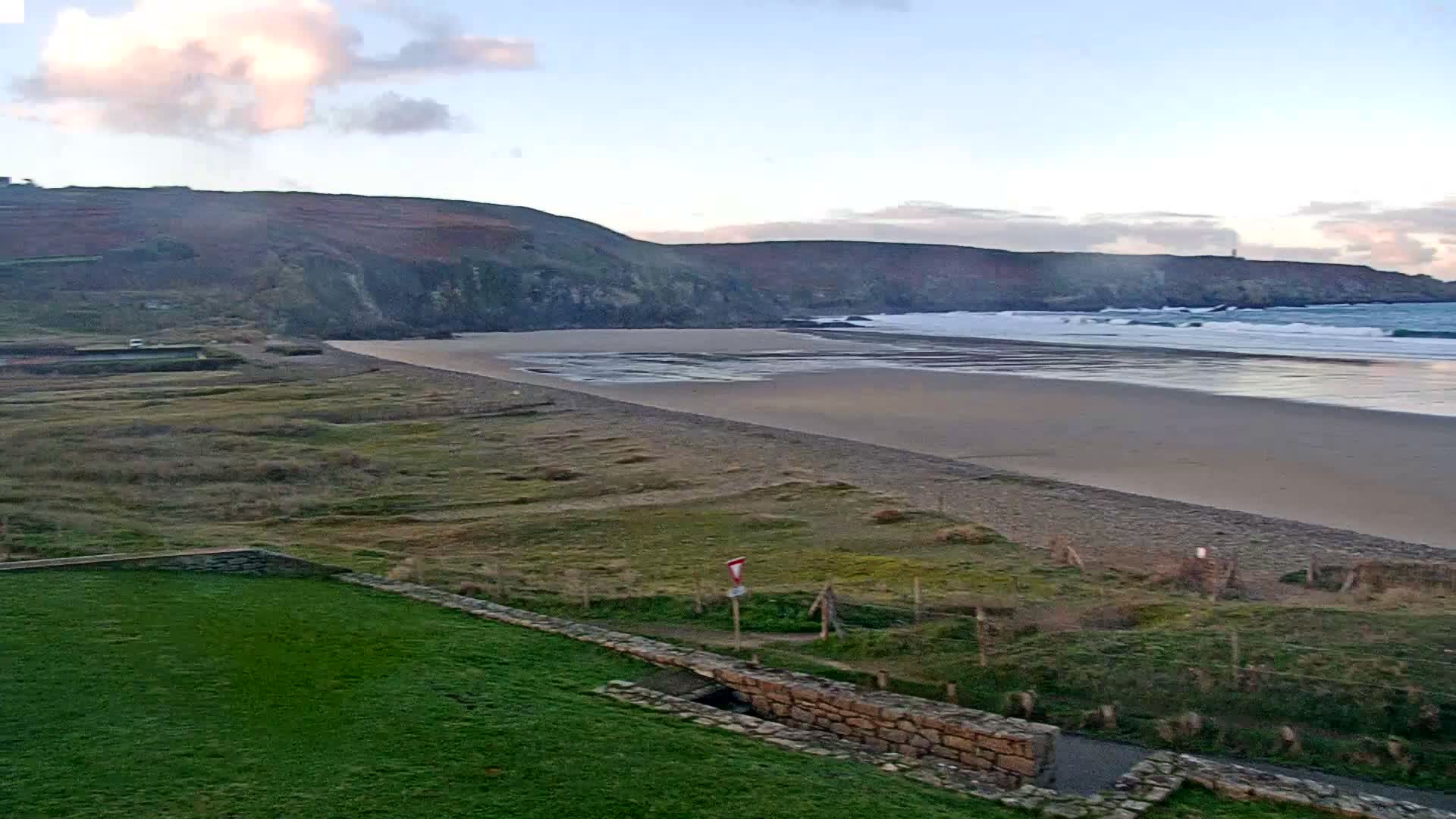 A wide coastal landscape is depicted under a partly cloudy sky, featuring a sandy beach with breaking waves, backed by rolling, vegetated cliffs, and a green grassy area with a stone wall in the foreground.
