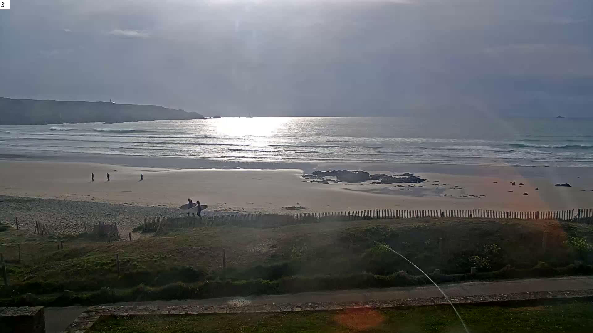 A wide coastal landscape is depicted under a partly cloudy sky, featuring a sandy beach with breaking waves, backed by rolling, vegetated cliffs, and a green grassy area with a stone wall in the foreground.