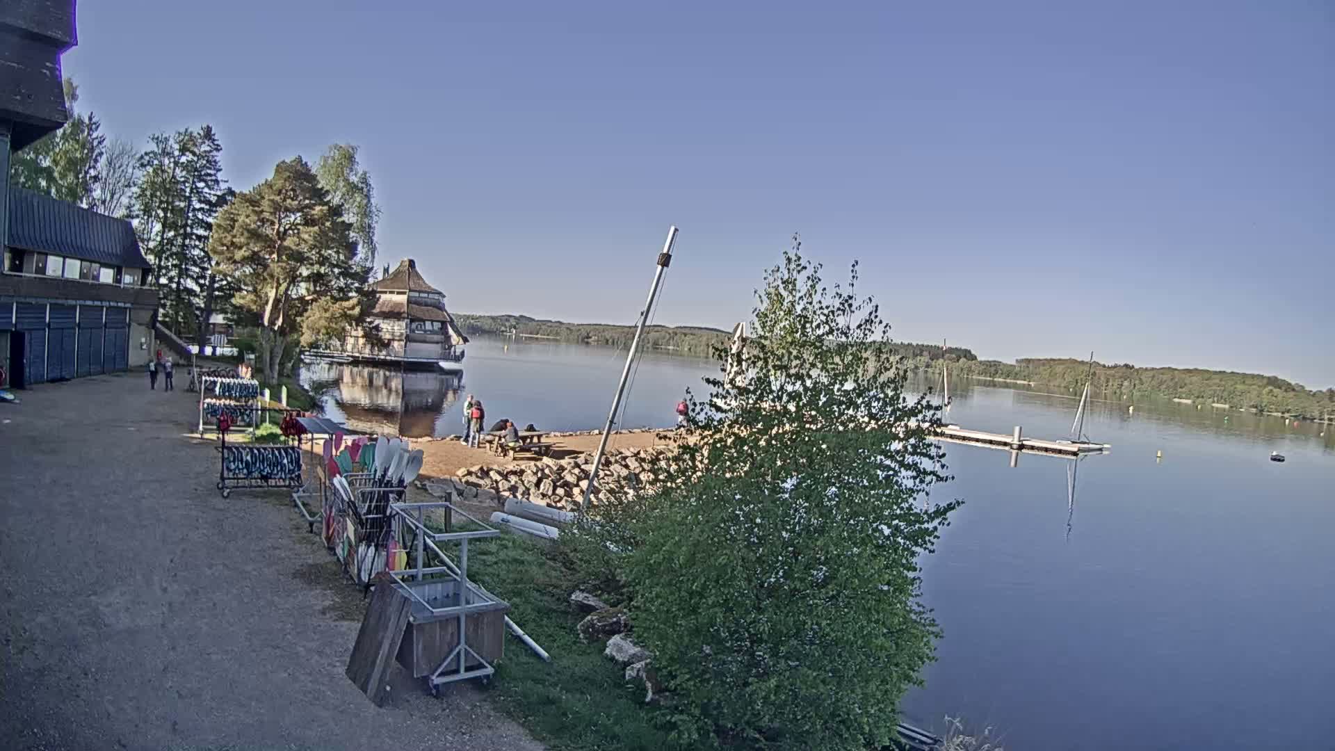 On a sunny day, a calm lake is seen with several people gathered near a building and a dock, with boats and paddles stored nearby.
