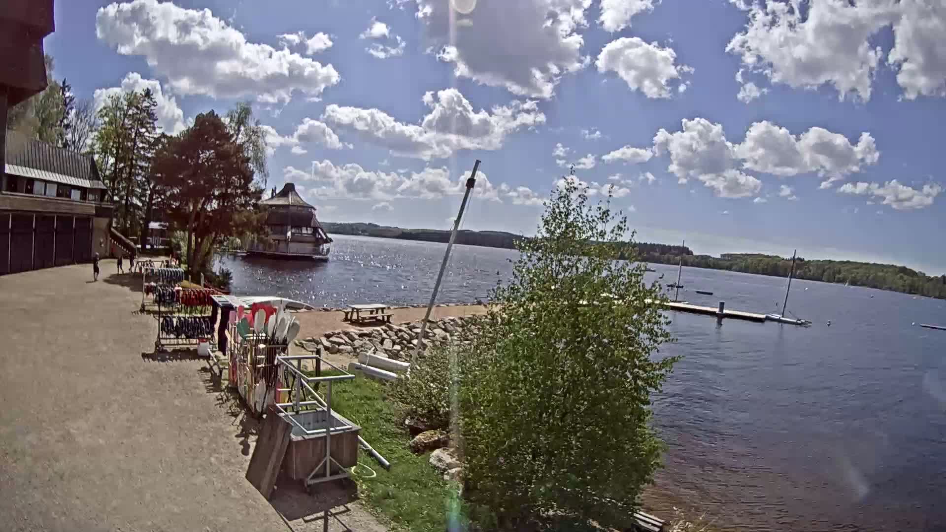 A lakeside area with a building, a dock with boats, and a rack of kayaks under a partly cloudy sky.