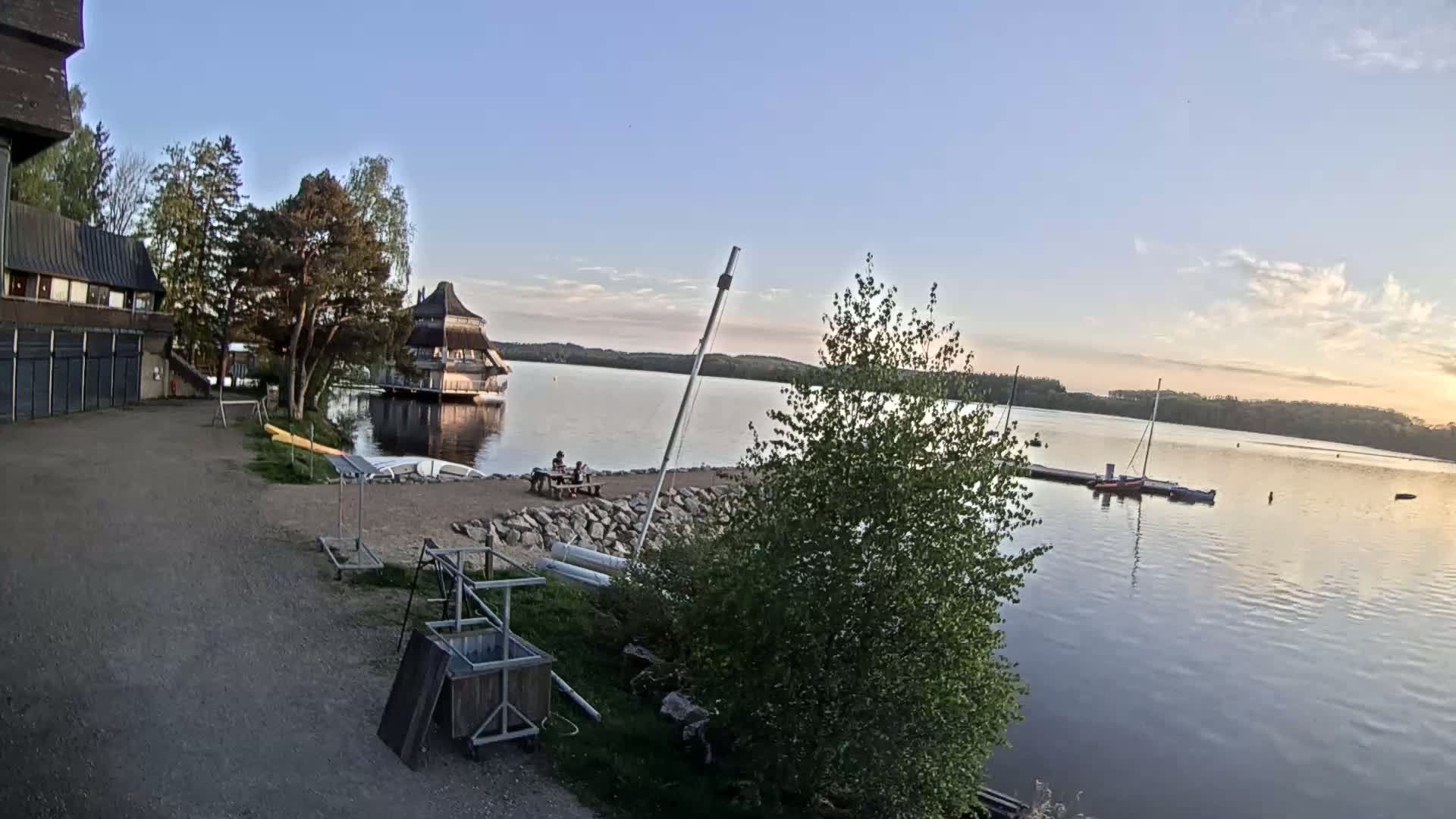A calm lake at sunset is viewed from a lakeside building, with a small pavilion visible on the water, a couple seated on a nearby bench, and boats docked at a pier.