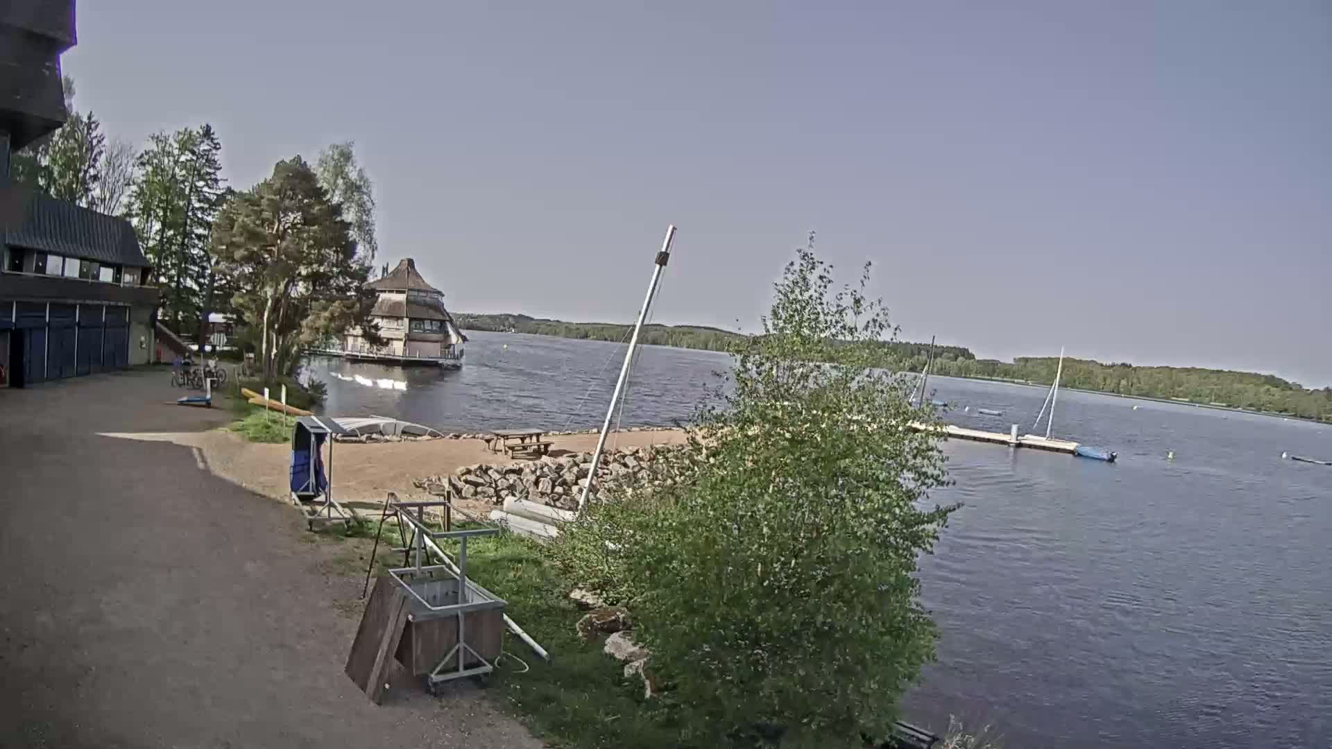 A lakeside scene on a partly sunny day shows a building, a small sandy beach with a picnic table, several boats on the water, and a grassy area with trees.
