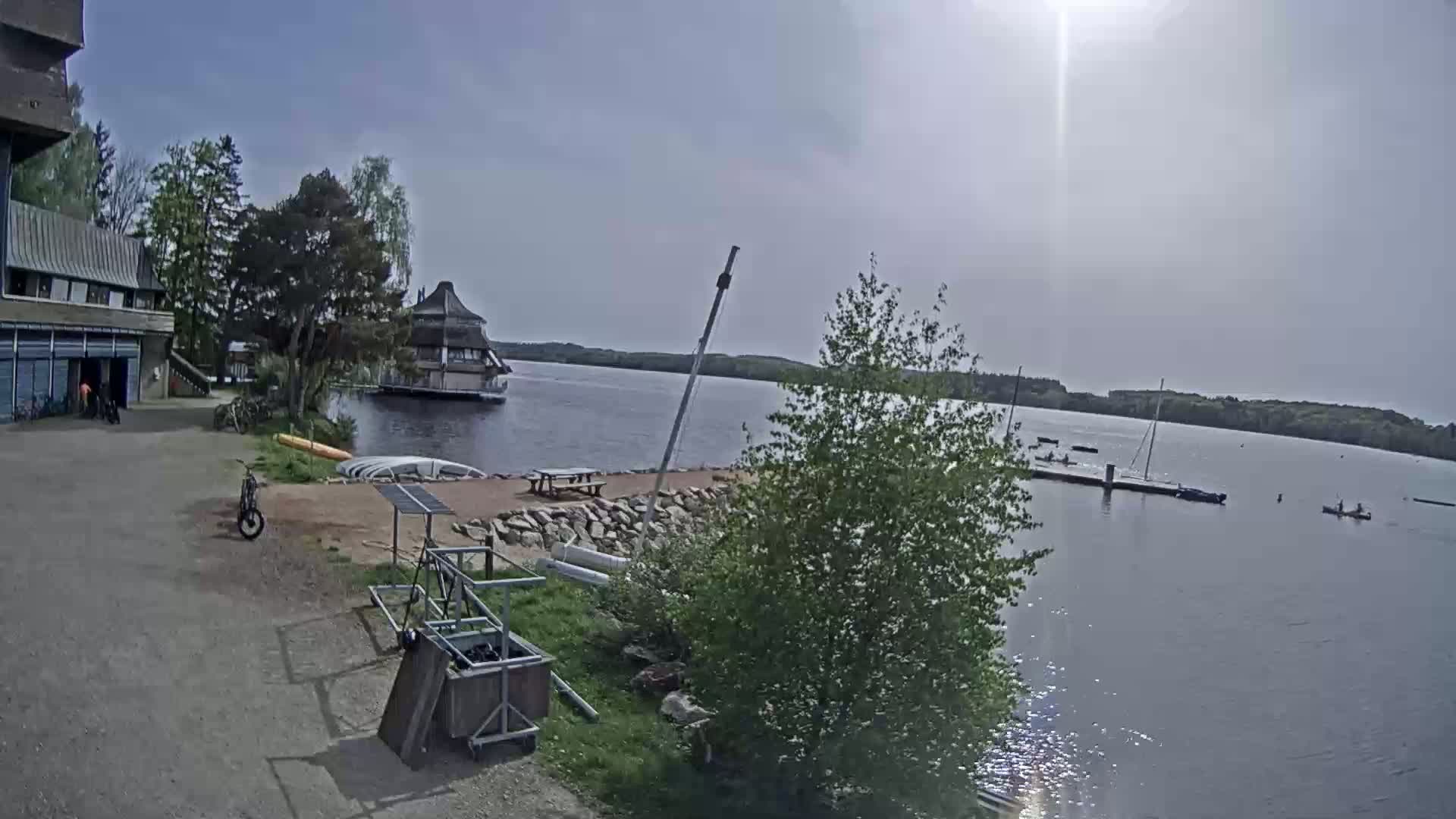 A sunny day at a lakeside area shows a building, boats on a dock, kayaks in the water, and bicycles near a paved area.