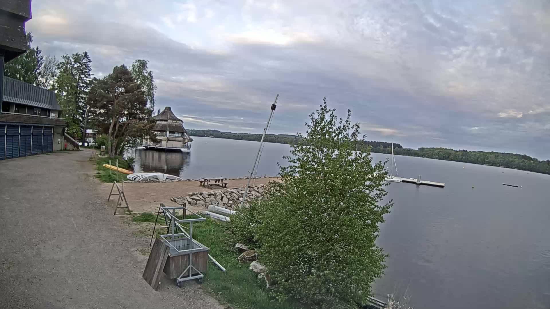 A calm lake under a cloudy sky is viewed from a lakeside area with a building, boats, and some trees.