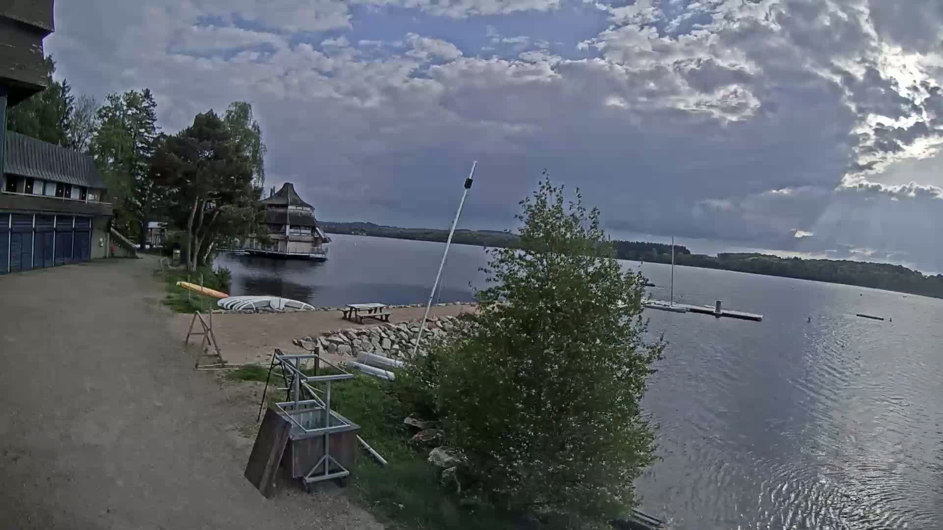 A lakeside view shows a building, boats, and a picnic table under a partly cloudy sky.