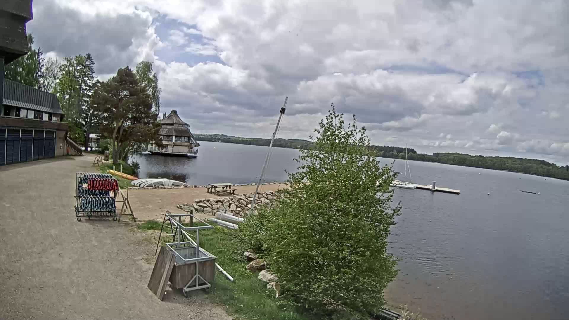 A lakeside view on a partly cloudy day shows a building, a small sandy beach with a picnic table, a boat dock, and several boats on the water.