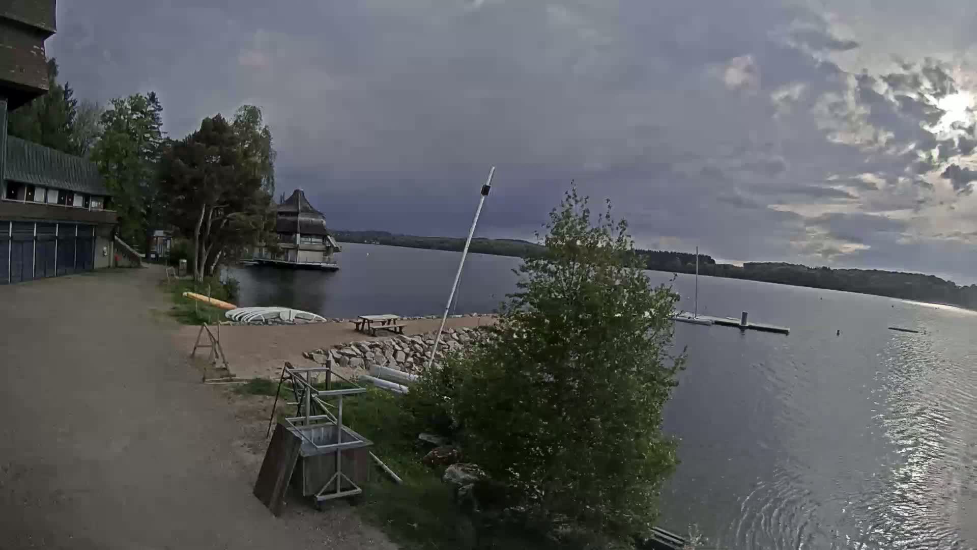 A calm lake under a cloudy sky is viewed from a lakeside area with buildings, a sandy beach, and several small boats.
