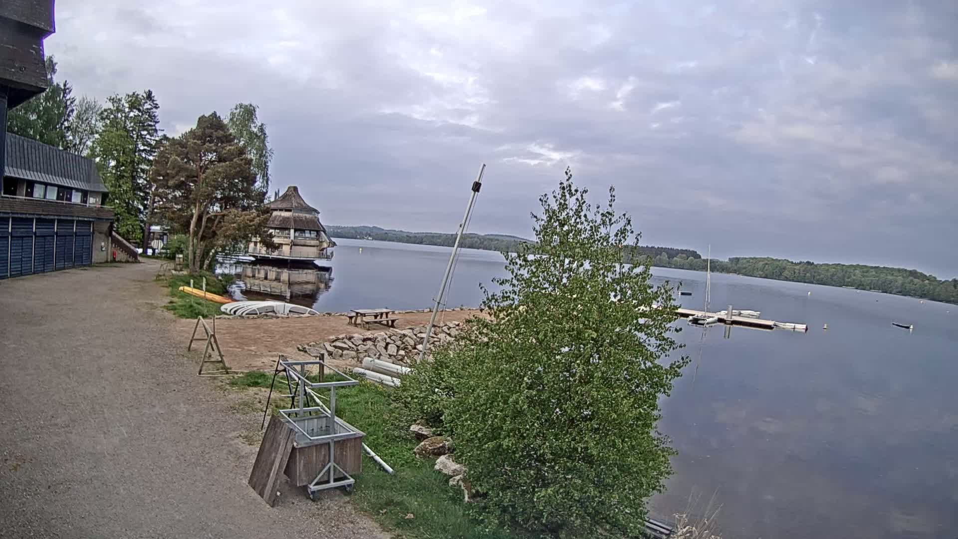 A calm lake is viewed from a lakeside area with a building, several small boats, and some trees under an overcast sky.