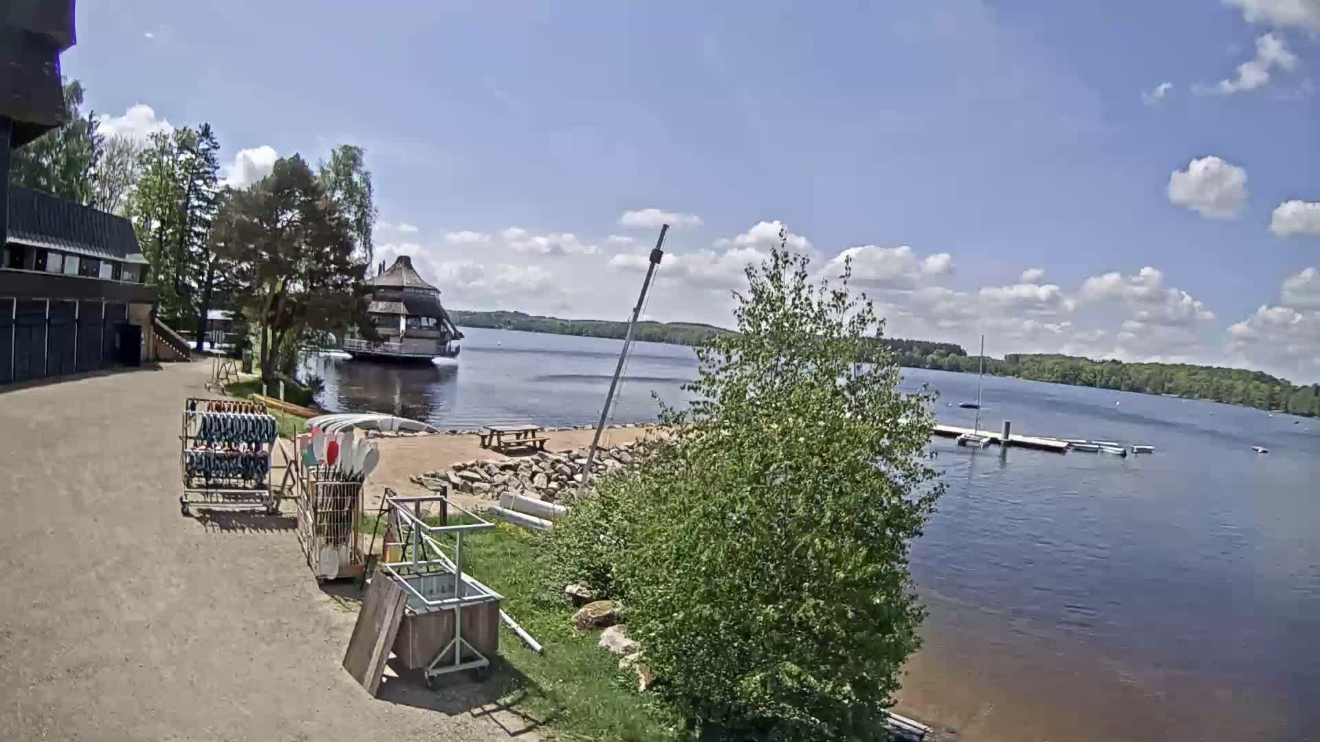 A lakeside scene on a partly sunny day shows a building, a dock with boats, and a rack of kayaks and paddles near a sandy shore.