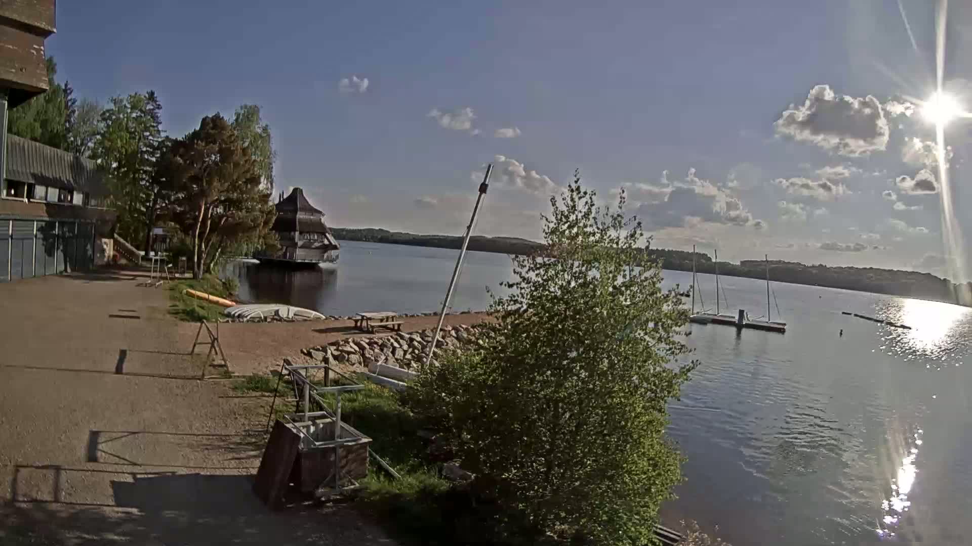 A sunny day at a lakeside area with a building, trees, several boats docked, and a picnic table.