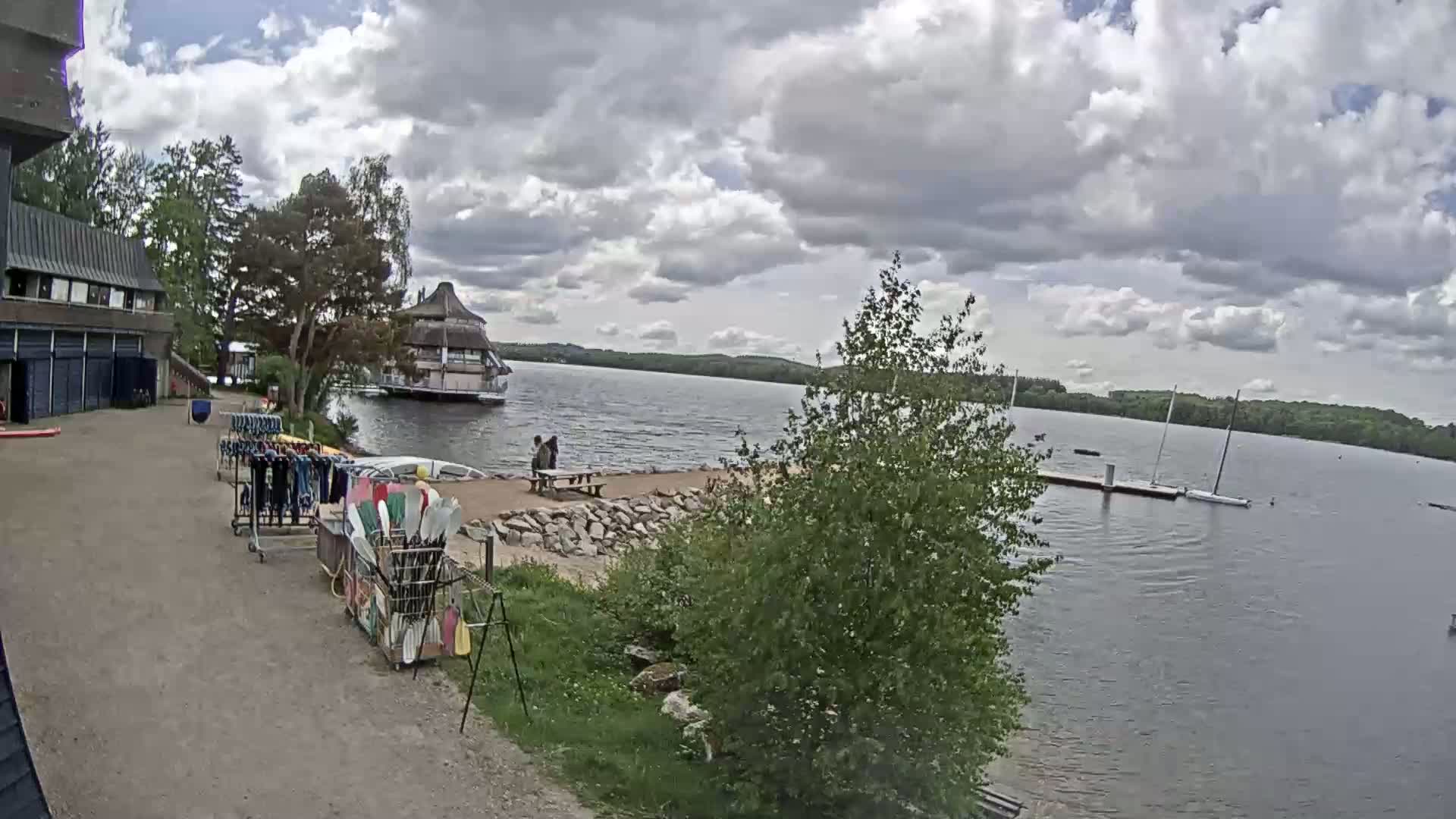 A lakeside scene under a partly cloudy sky shows a building, a small restaurant, a sandy beach with picnic tables, racks of water sports equipment, and sailboats moored on a dock.
