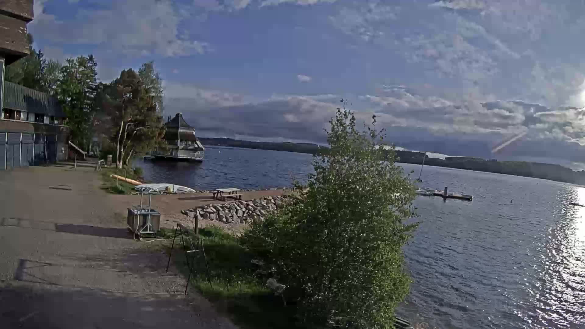A partly sunny day shows a lakeside view with a building, a small dock, and picnic tables near a rocky shoreline.