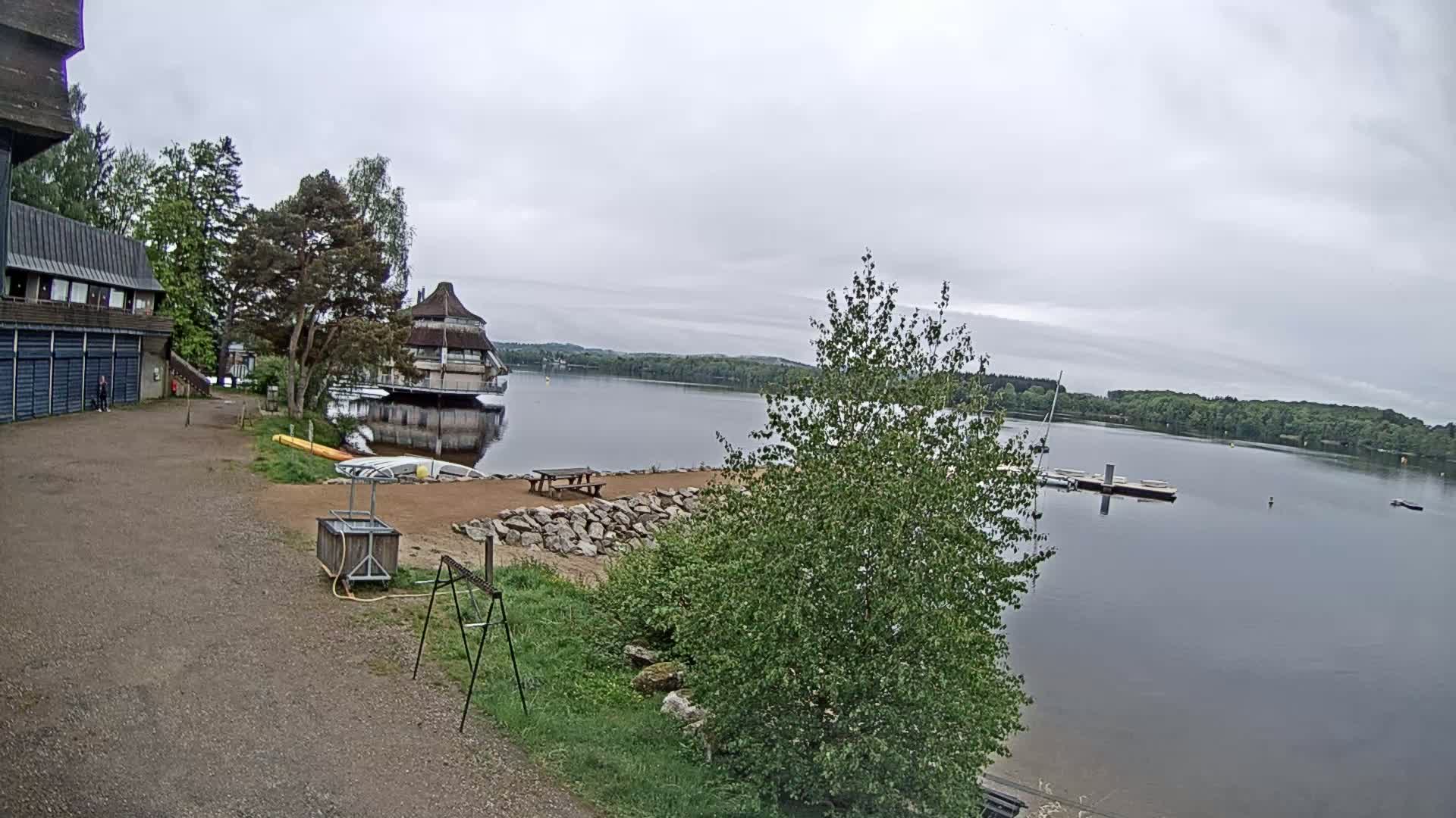 A lakeside view on an overcast day shows a building, a sandy area with a picnic table, and a calm lake with a dock and small boats.