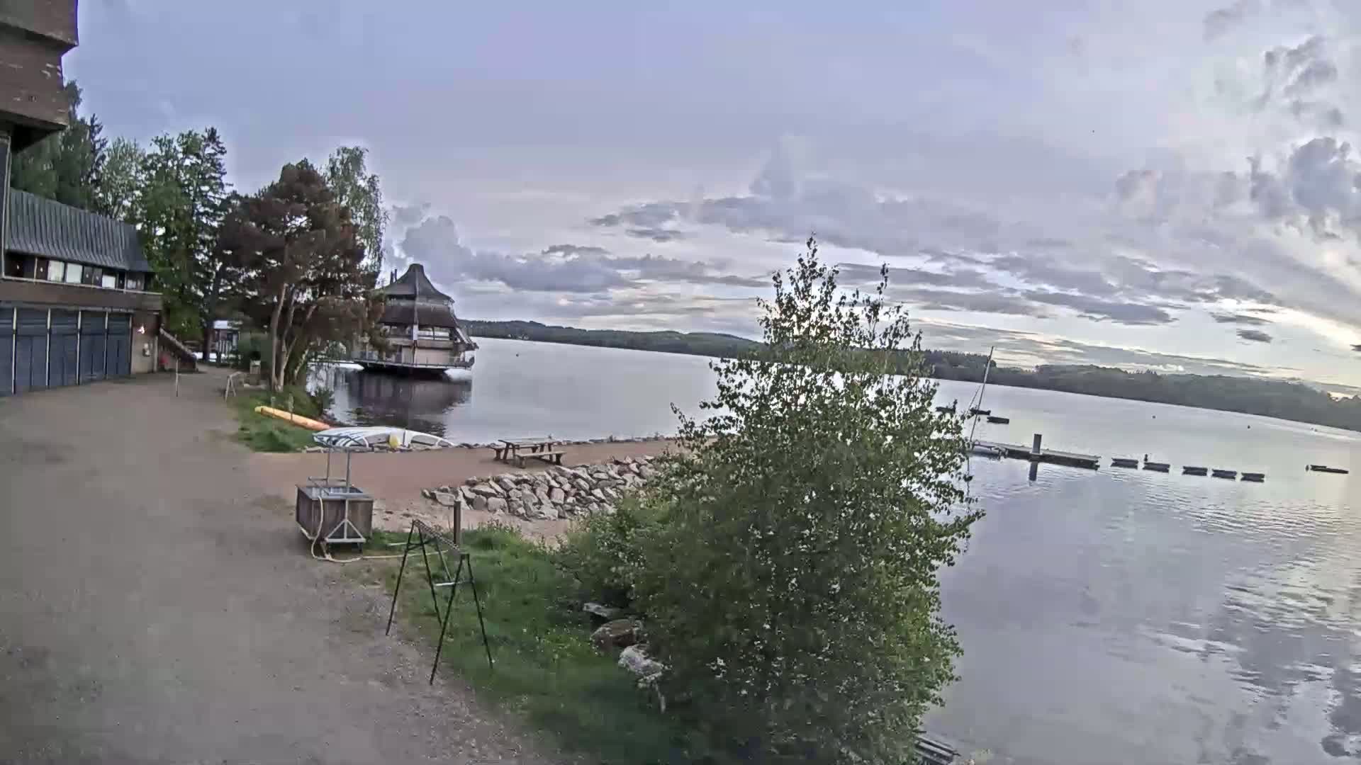 A calm lake is seen under a cloudy sky, with a building, small beach area, and a floating structure on the water's edge.