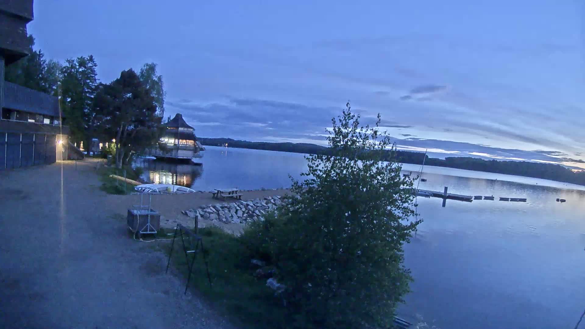 A lakeside view at dusk shows a building, a small beach area, and a calm lake with a dock.