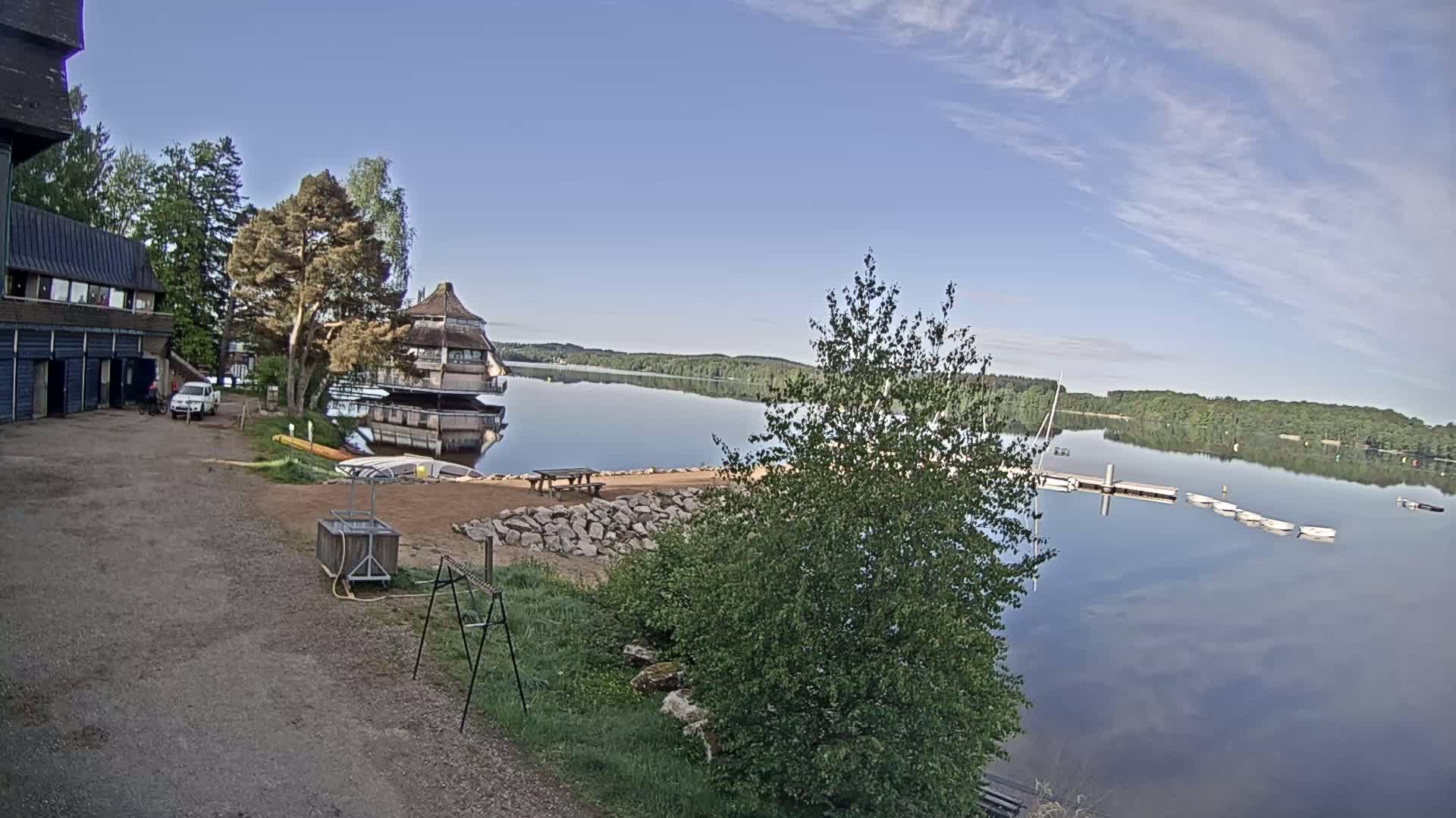 A calm lake reflects a clear blue sky, with trees, a building, and a small beach area visible along the shoreline.