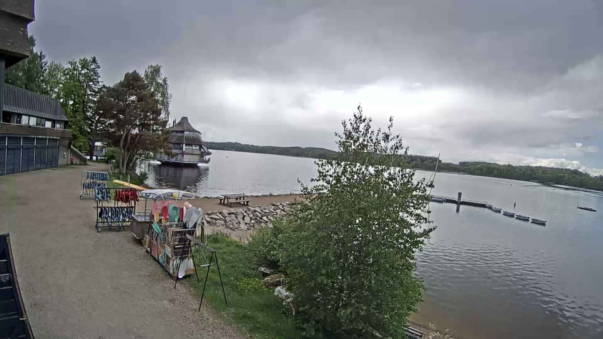 Kayaks and other water equipment are stored near a building overlooking a calm lake under an overcast sky.