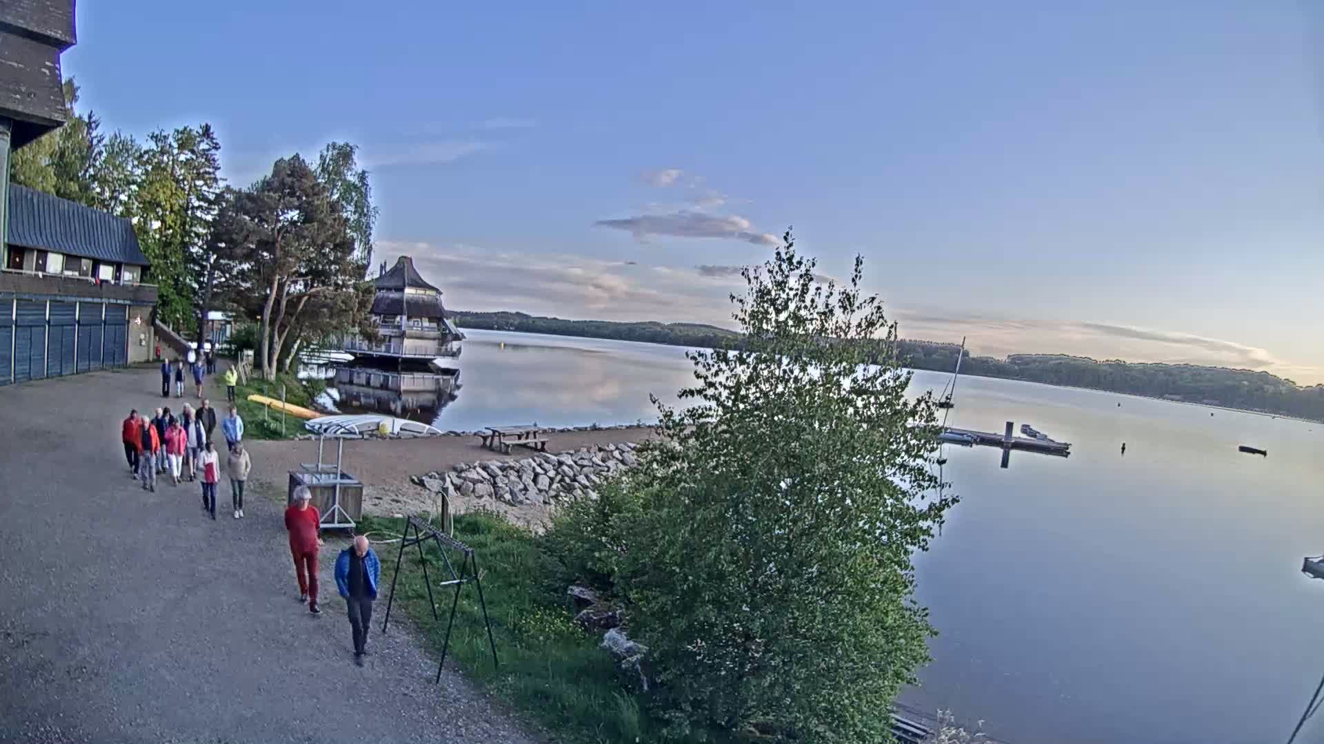 A group of people walk along a paved path next to a calm lake under a mostly clear, light blue sky, with a building and trees visible in the background.