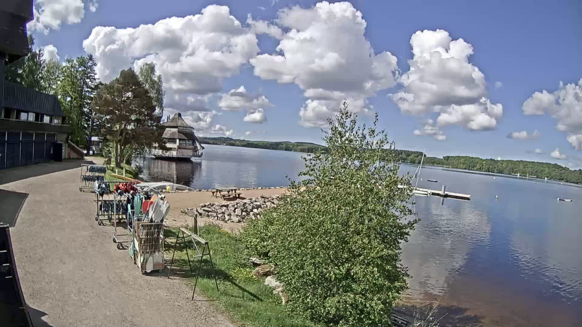 A lakeside view on a partly sunny day shows a calm lake with a building on the shore, a small beach area, and equipment carts with water sports gear.