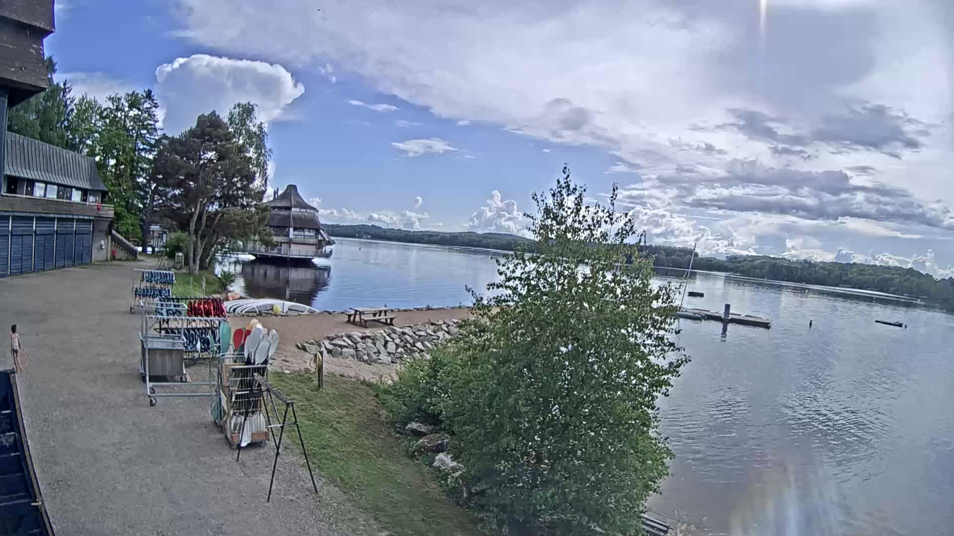 A partly cloudy day shows a calm lake with a building on the shore,  kayaks and other watercraft stored nearby, and trees surrounding the waterfront.
