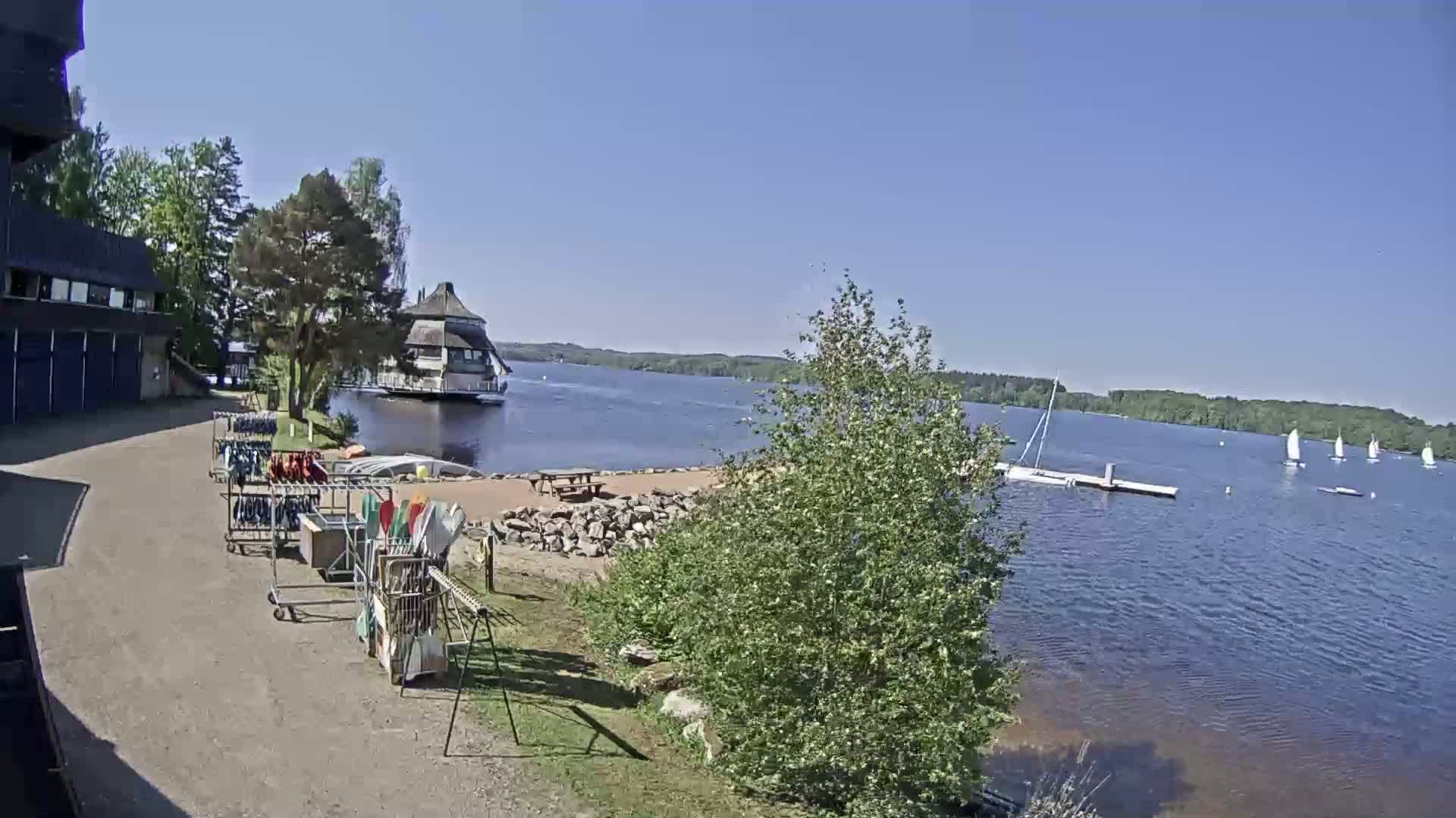 A lakeside view on a sunny day shows a building, equipment racks, a picnic table, sailboats on the water, and lush green trees.