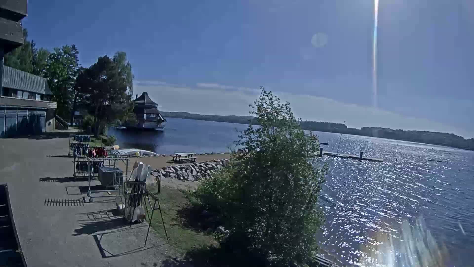 A sunny day shows a calm lake with a building on the far shore, equipment racks near a building and small beach on the near shore, and some trees and vegetation.