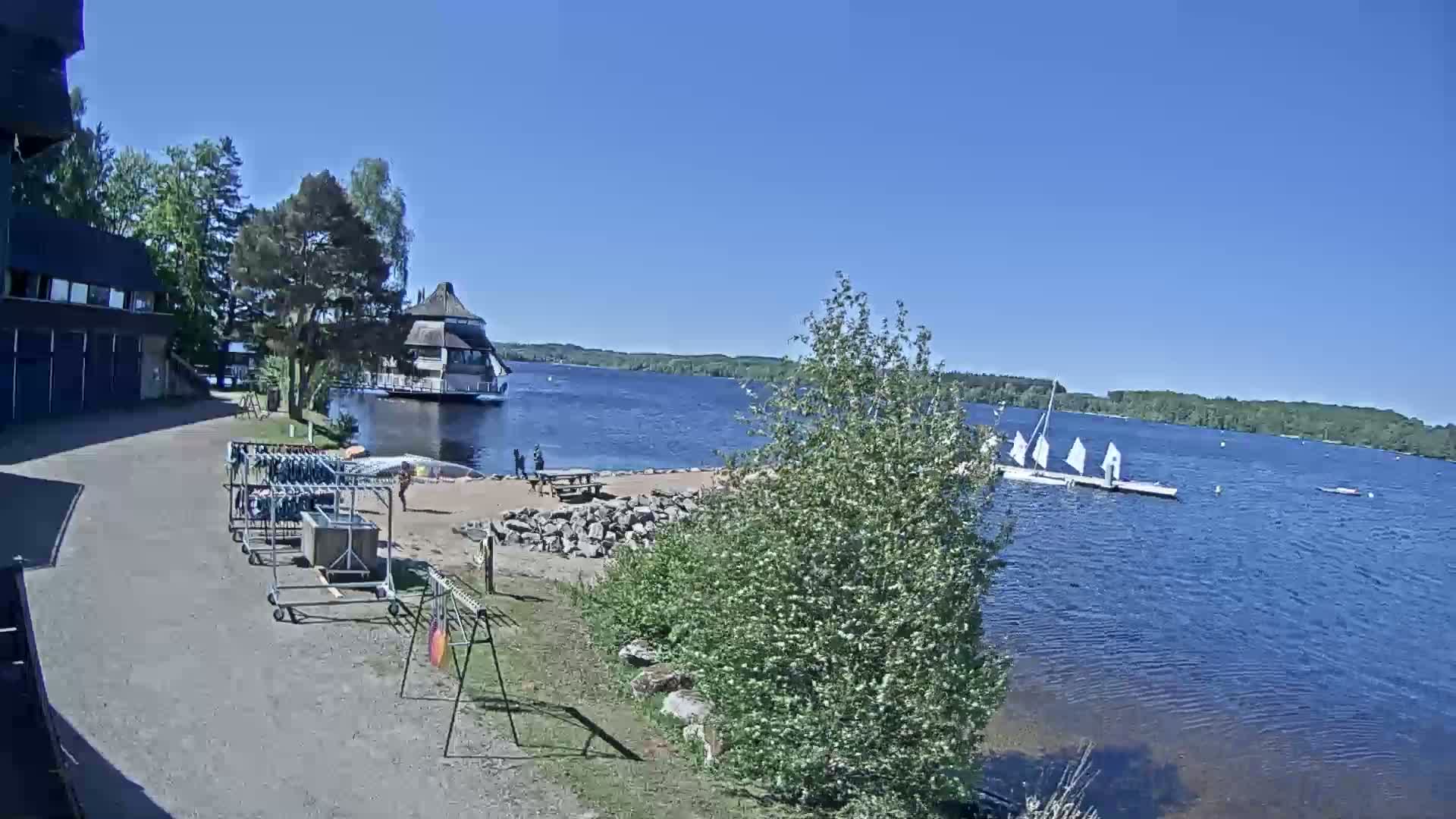 A sunny day at a lakeside recreational area shows a sandy beach with picnic tables, several sailboats on the water, and equipment racks near a building.
