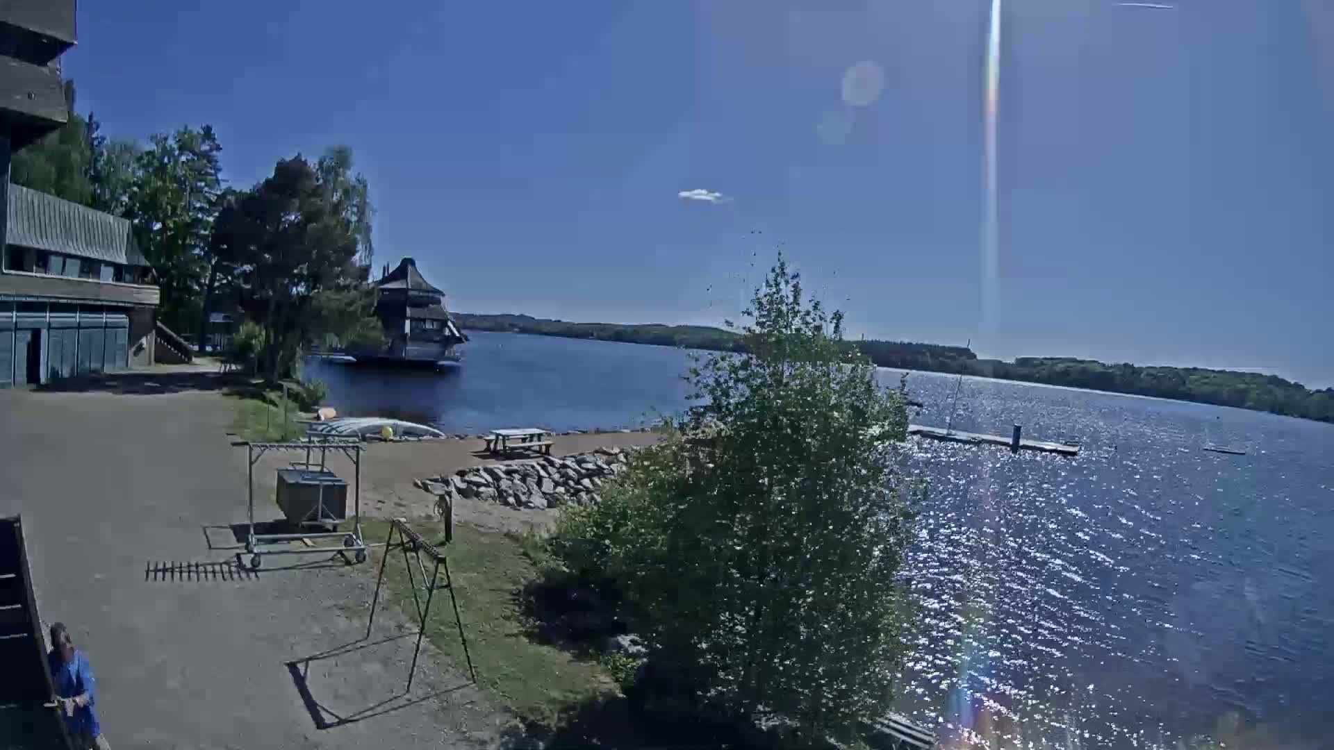 A sunny day at a lakeside facility shows a building, a dock, and various outdoor equipment, with a calm lake in the background.