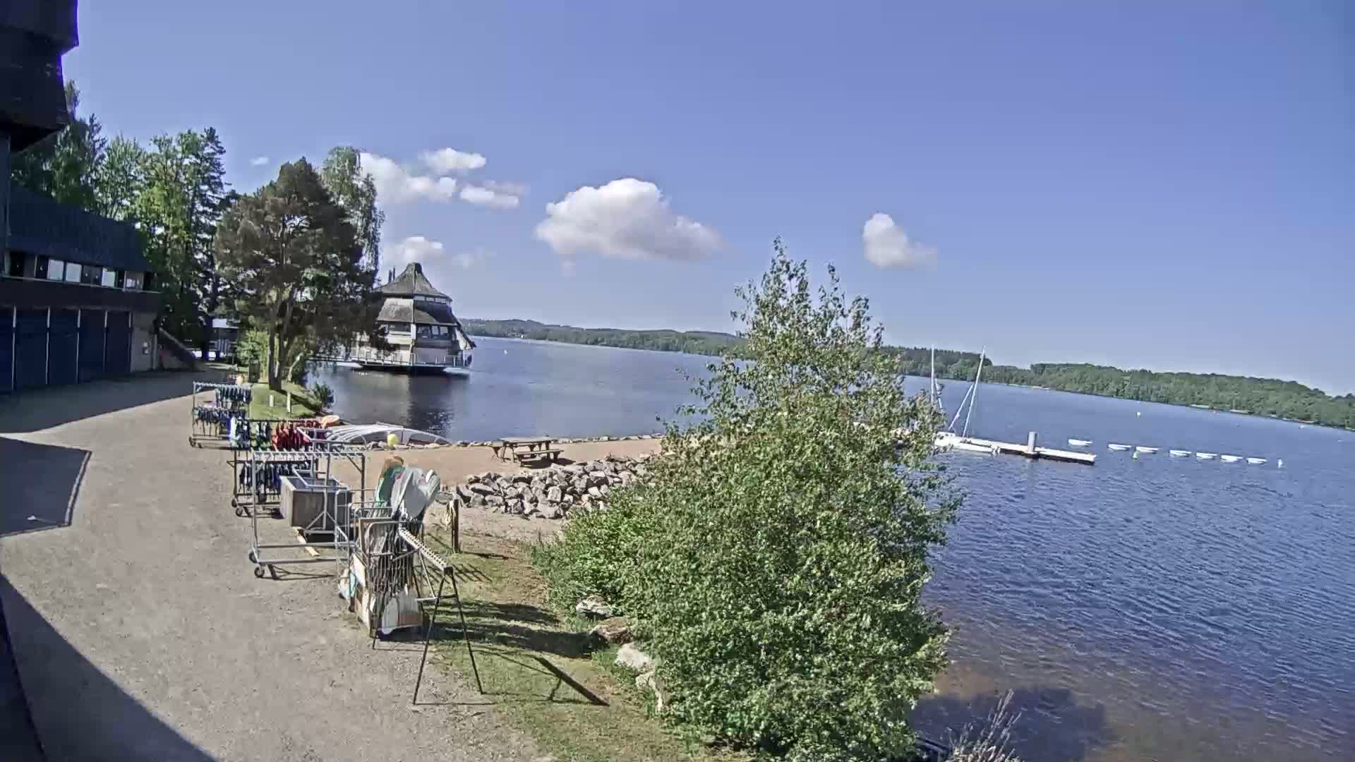 A lakeside view on a sunny day shows a building, equipment racks, a sandy area with a picnic table, and several small boats moored on a dock.