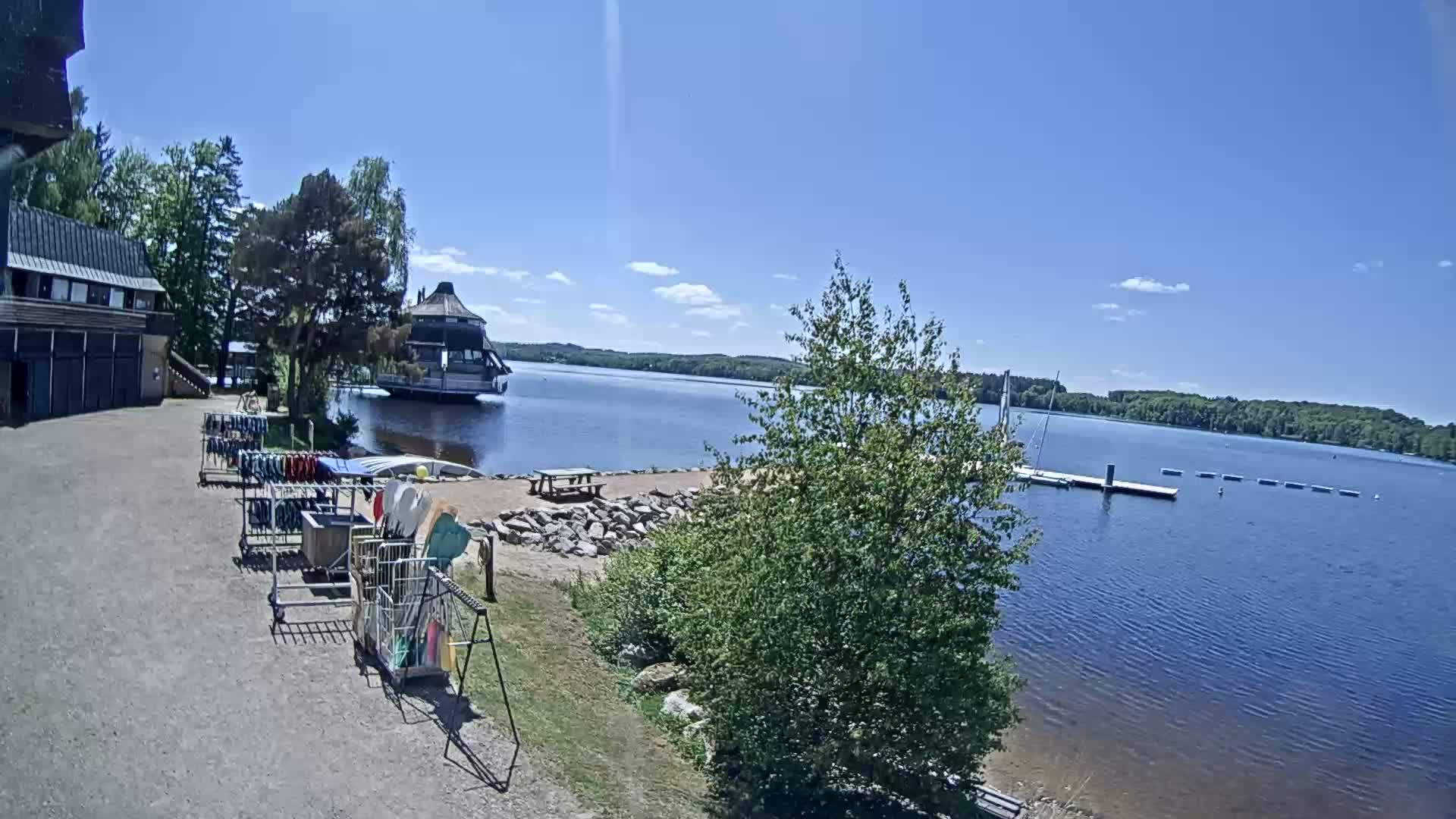A lakeside scene on a sunny day shows a building with equipment racks, a floating structure on the water, and a dock in the distance.