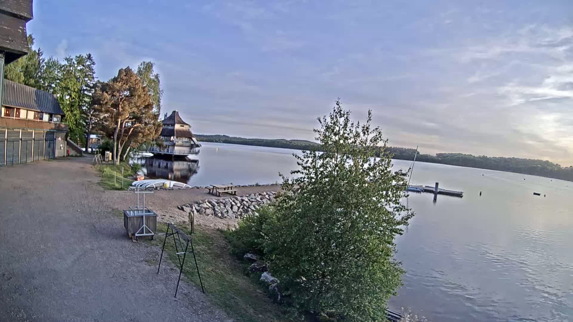 A calm lake is seen under a mostly clear sky, with a building and a small dock visible along the shoreline, surrounded by trees and some outdoor furniture.
